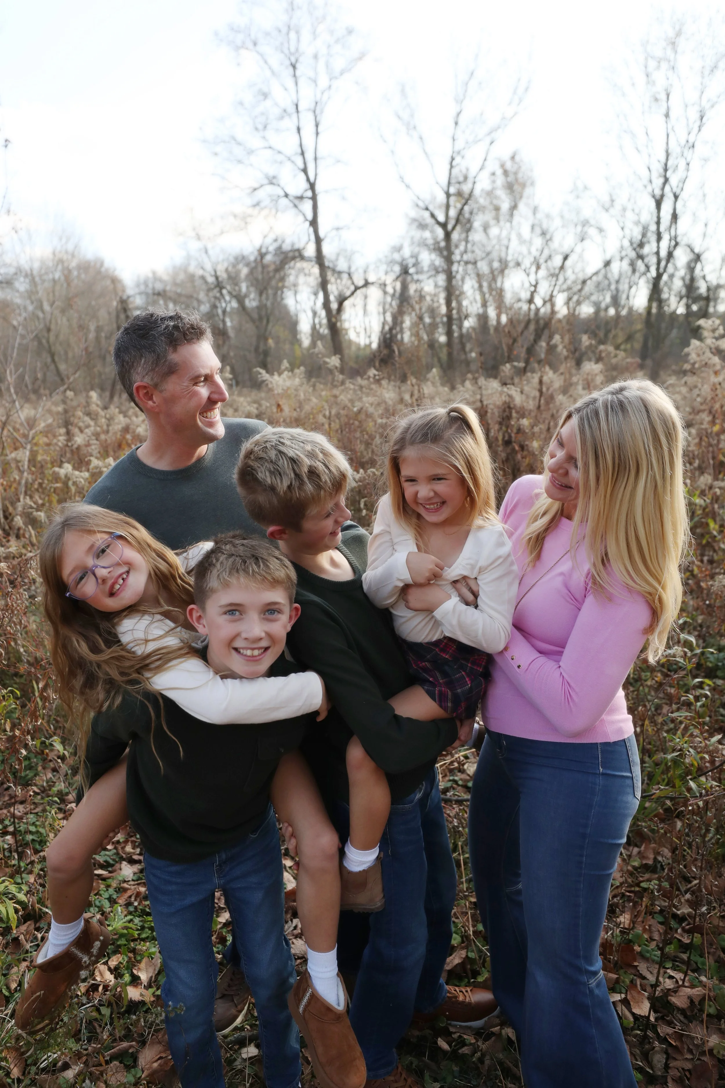 Family with four children laughing and hugging outdoors in a wooded area during fall, dressed casually.