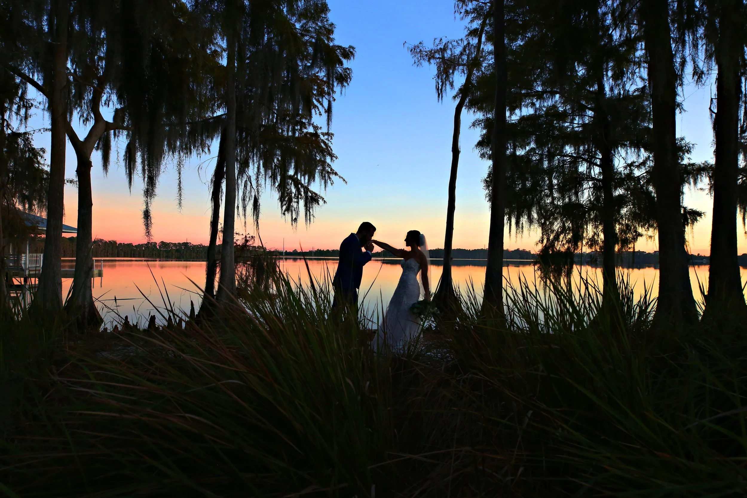 Silhouette of a newlywed couple by a lake at sunset, surrounded by trees.