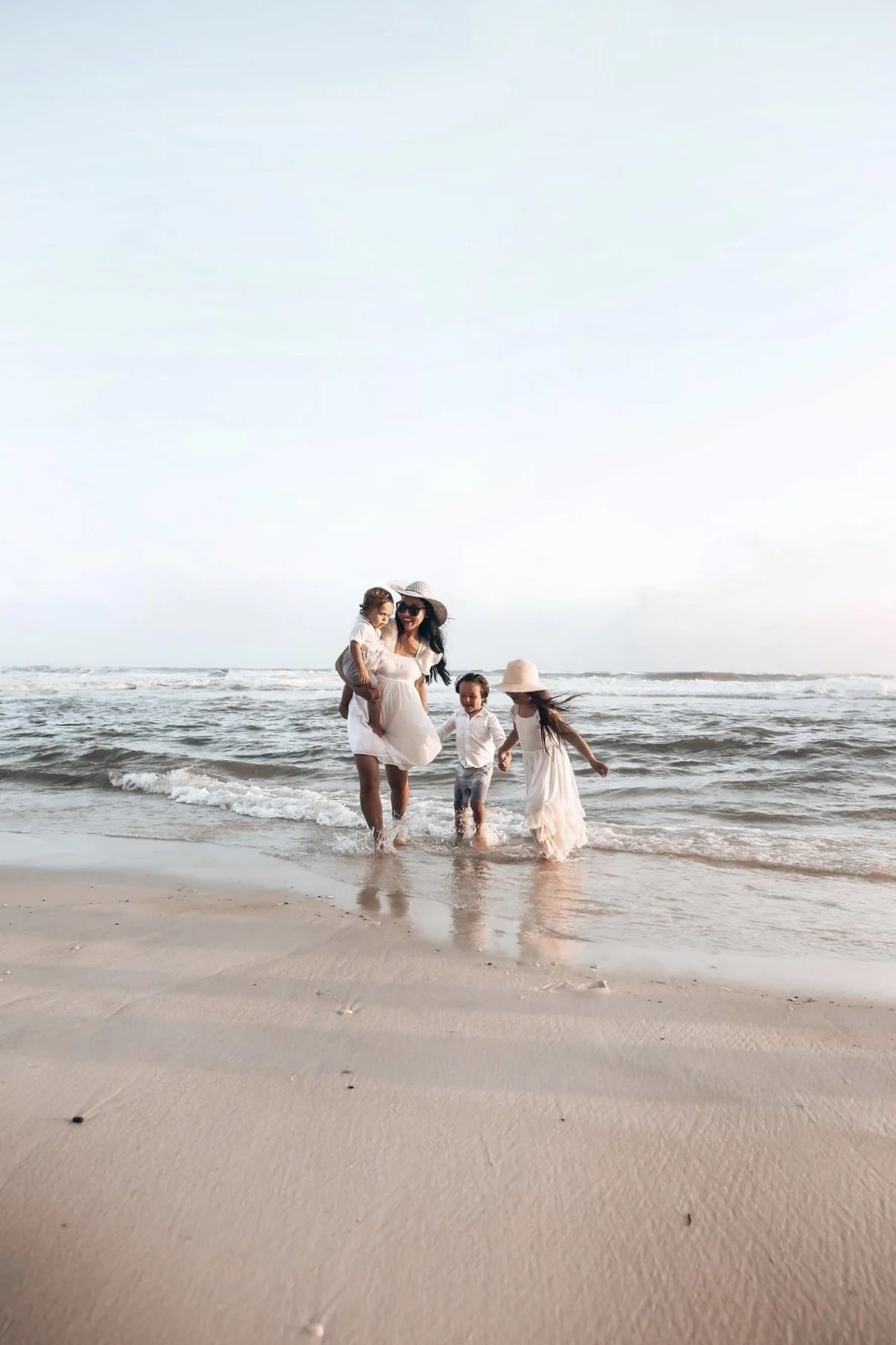 A woman and three children walking and playing in the shallow water at the beach, all dressed in white and wearing sun hats.