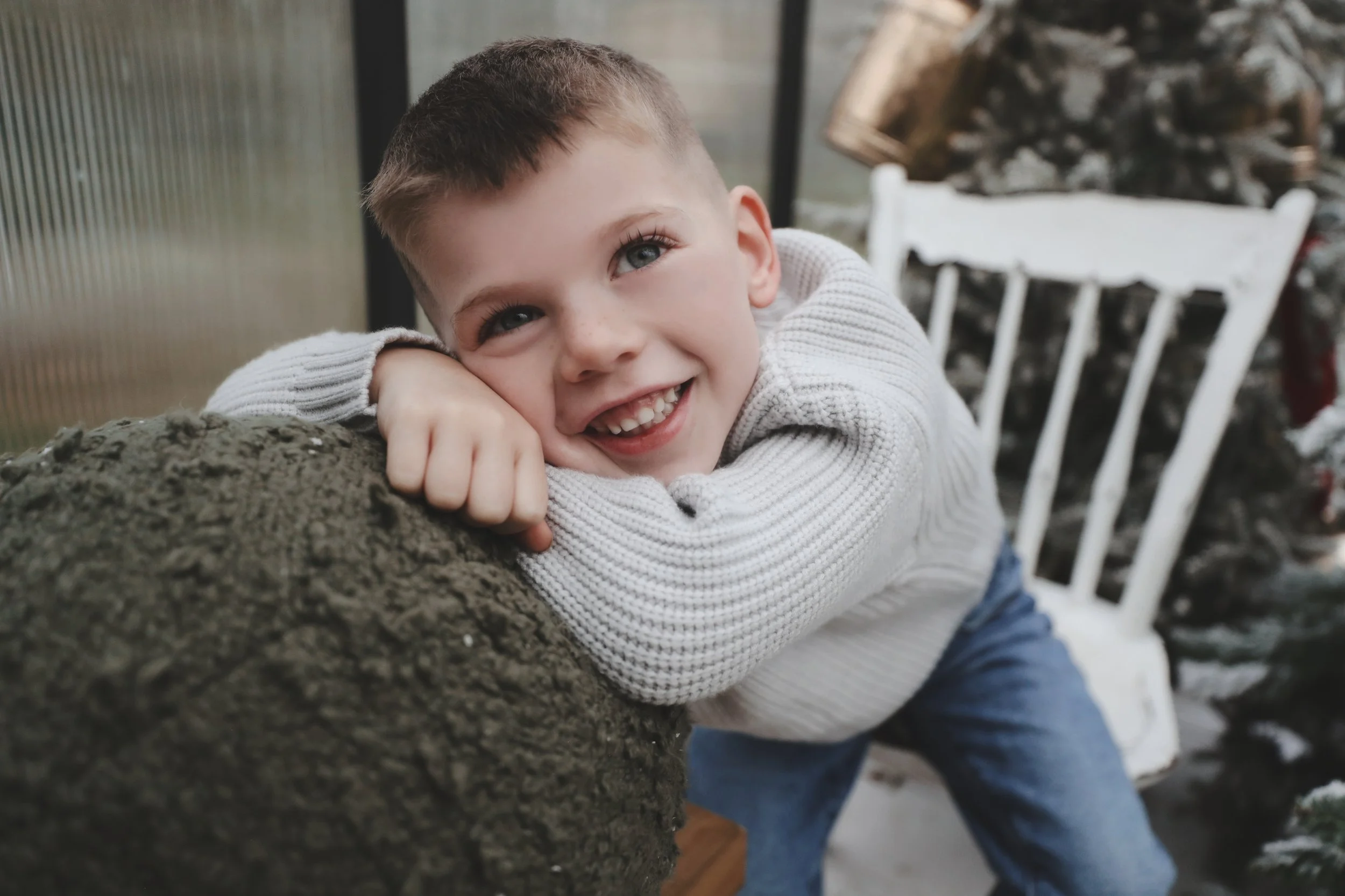 Happy young boy with short brown hair, blue eyes, and a wide smile showing missing front teeth, resting on a large log outdoors. He is wearing a light gray sweater and blue jeans.