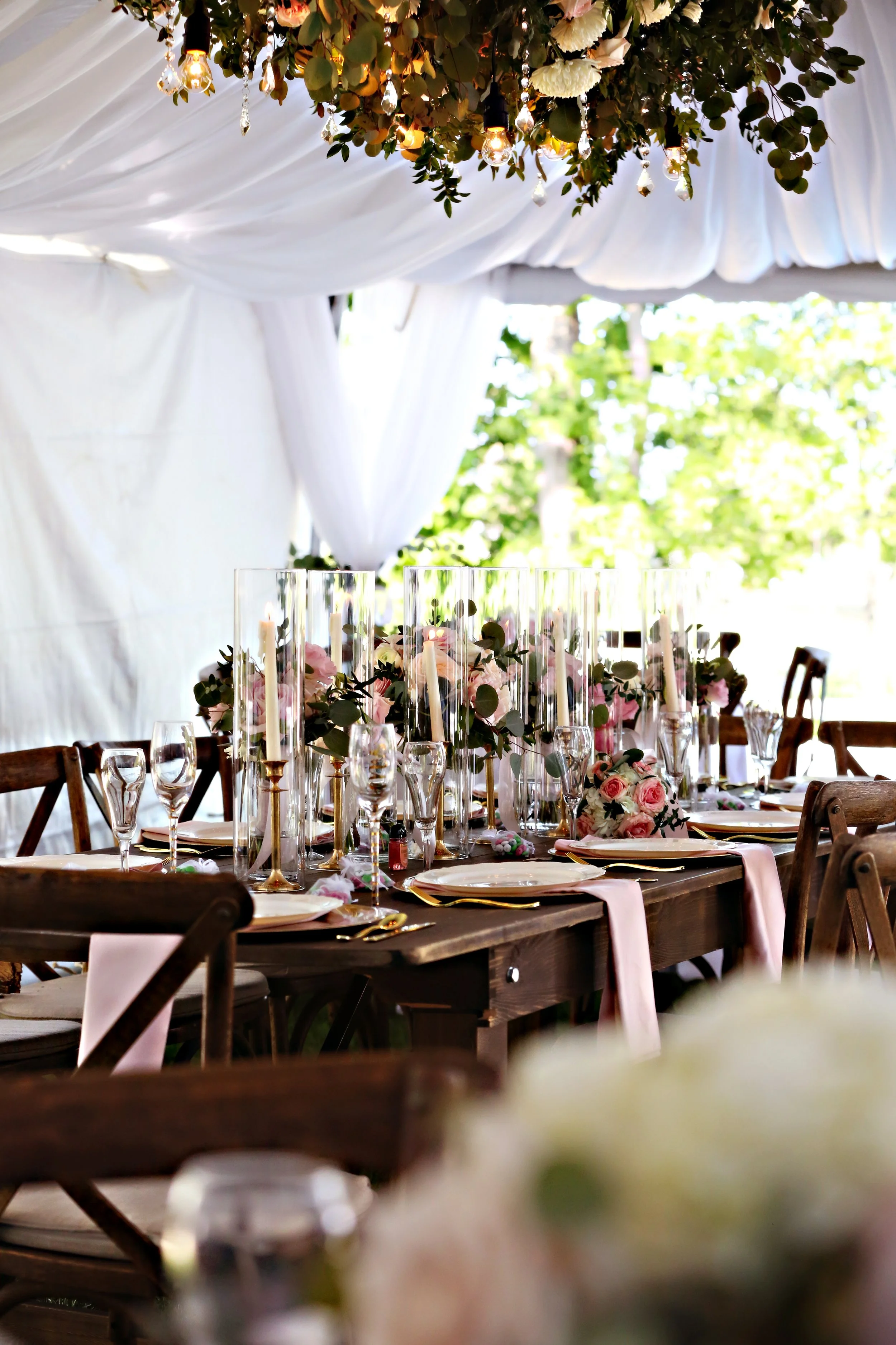 Elegant wedding reception table with pink and white flowers, tall glass candle holders, wine glasses, and pink napkins in a tent with white draping and greenery outside.