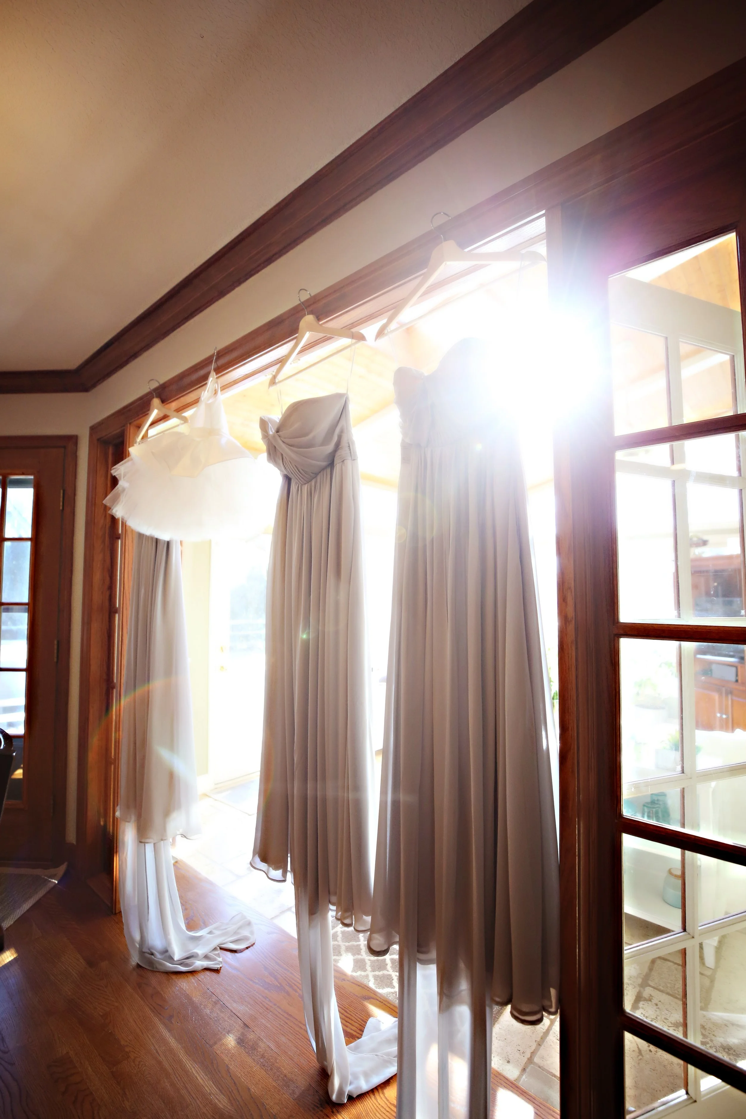 Four white dresses hanging on hangers in front of a sliding glass door, with sunlight streaming in.