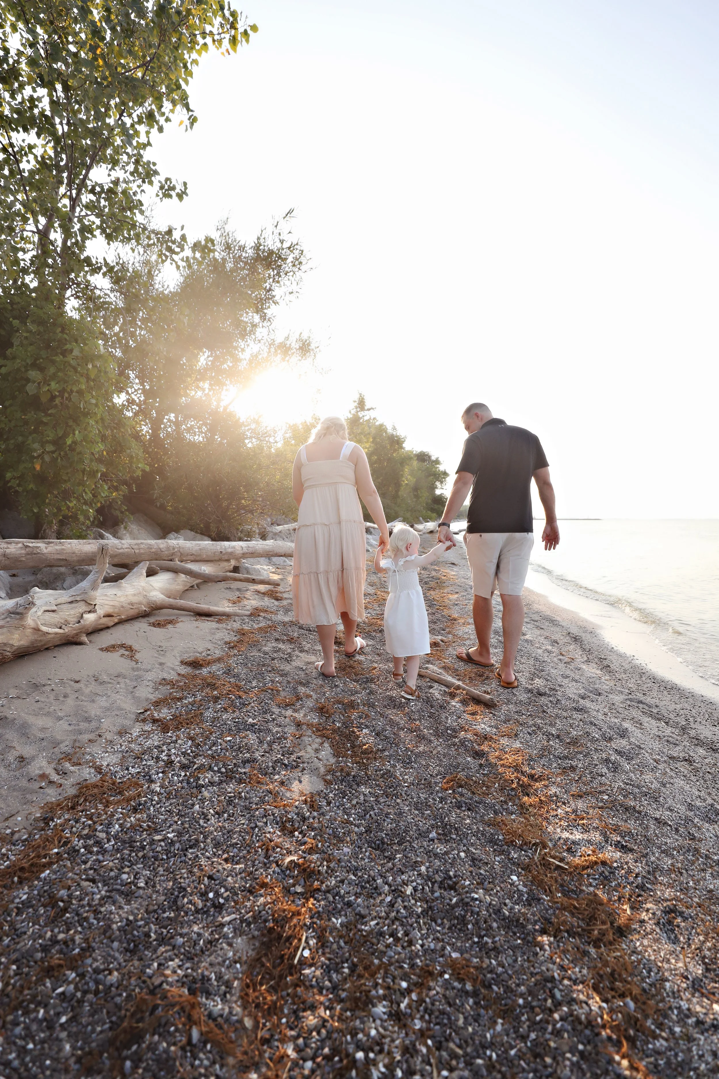 A family of three, a man, a woman, and a young girl, walking hand in hand along a sandy and pebbly beach at sunset, with trees on the left and the ocean on the right.