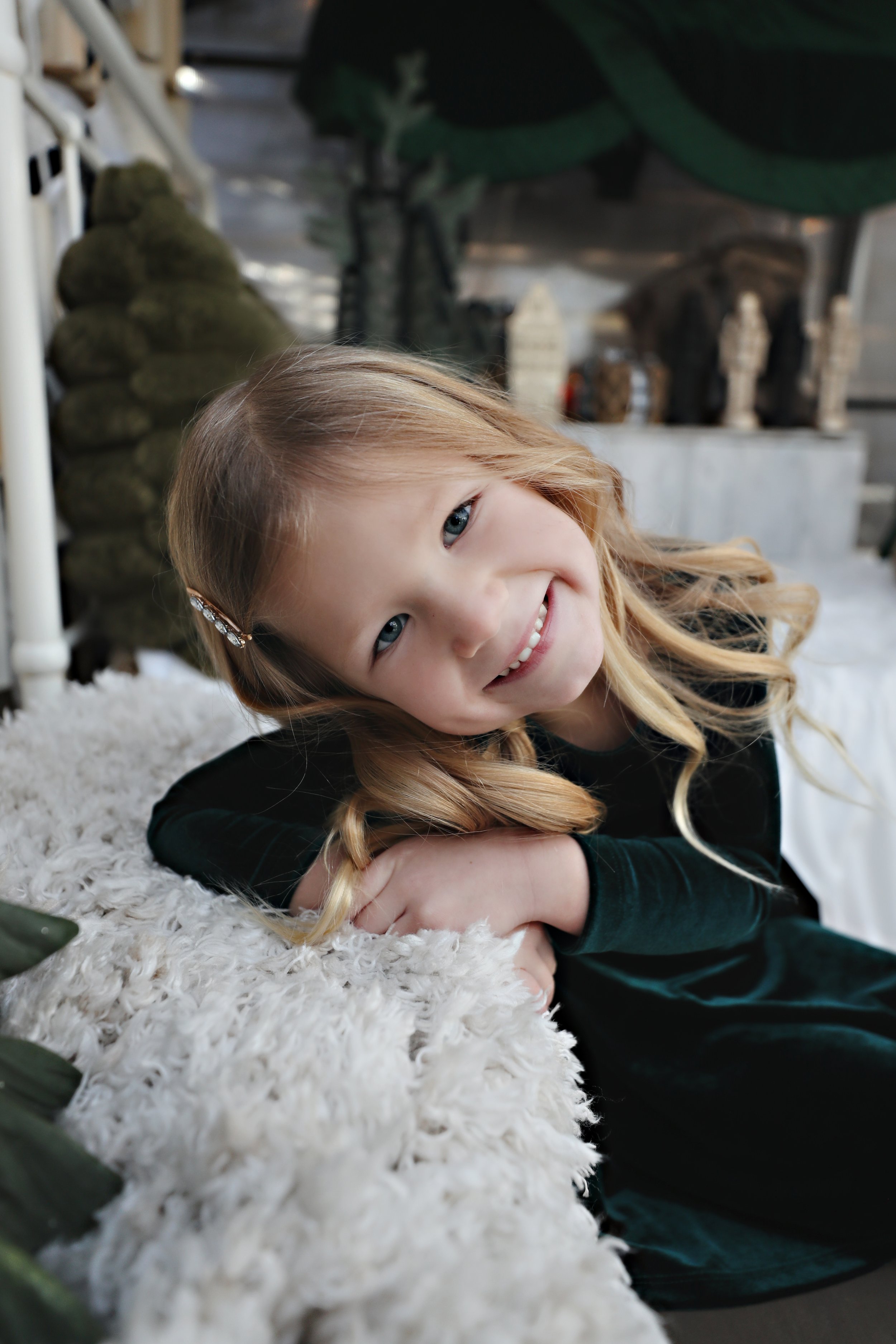 A young girl with blonde curly hair and a decorative hair clip, smiling and resting her head on her arms on a fluffy white surface.
