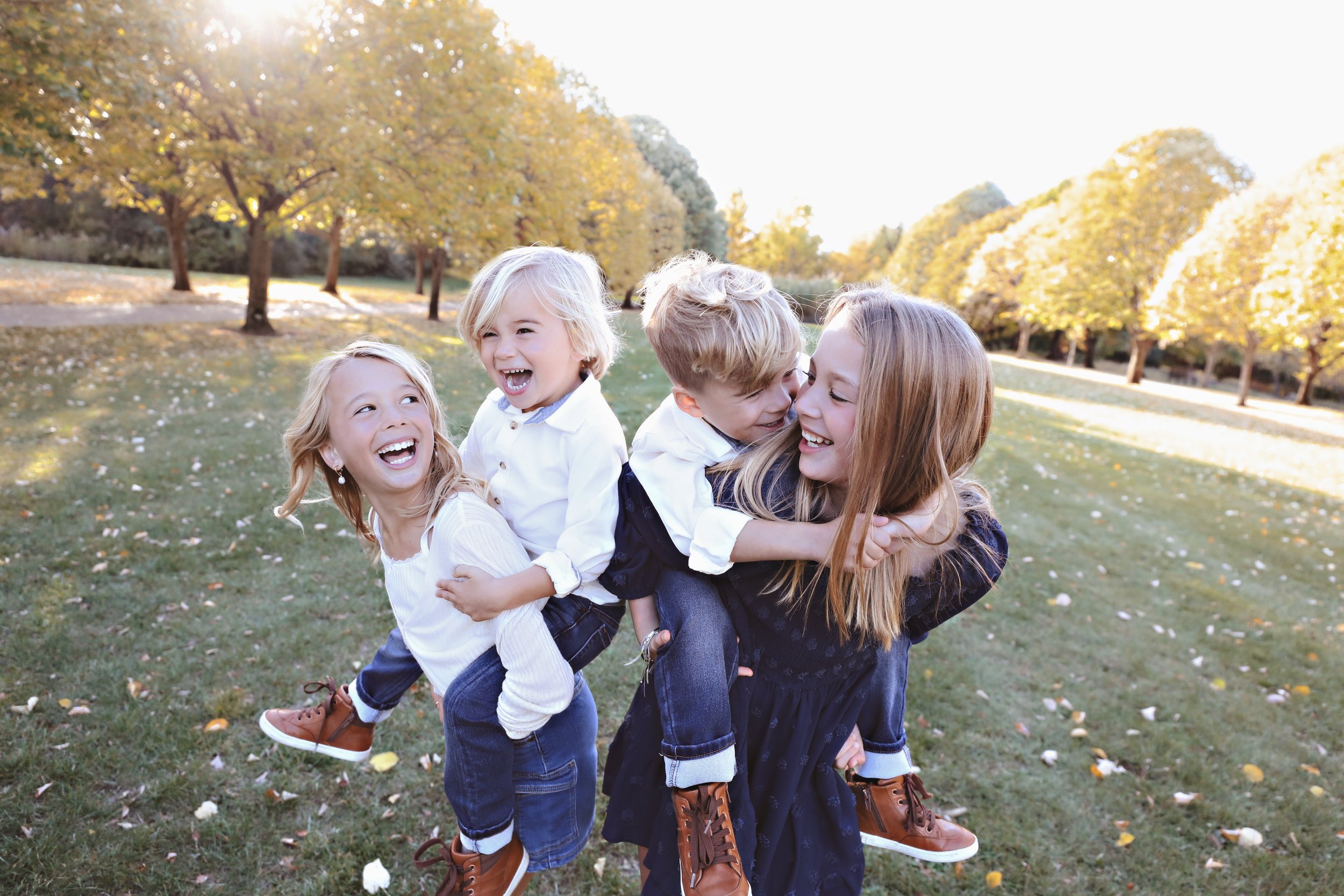 A woman giving piggyback rides to three children in a park with trees and autumn foliage, all smiling and laughing.