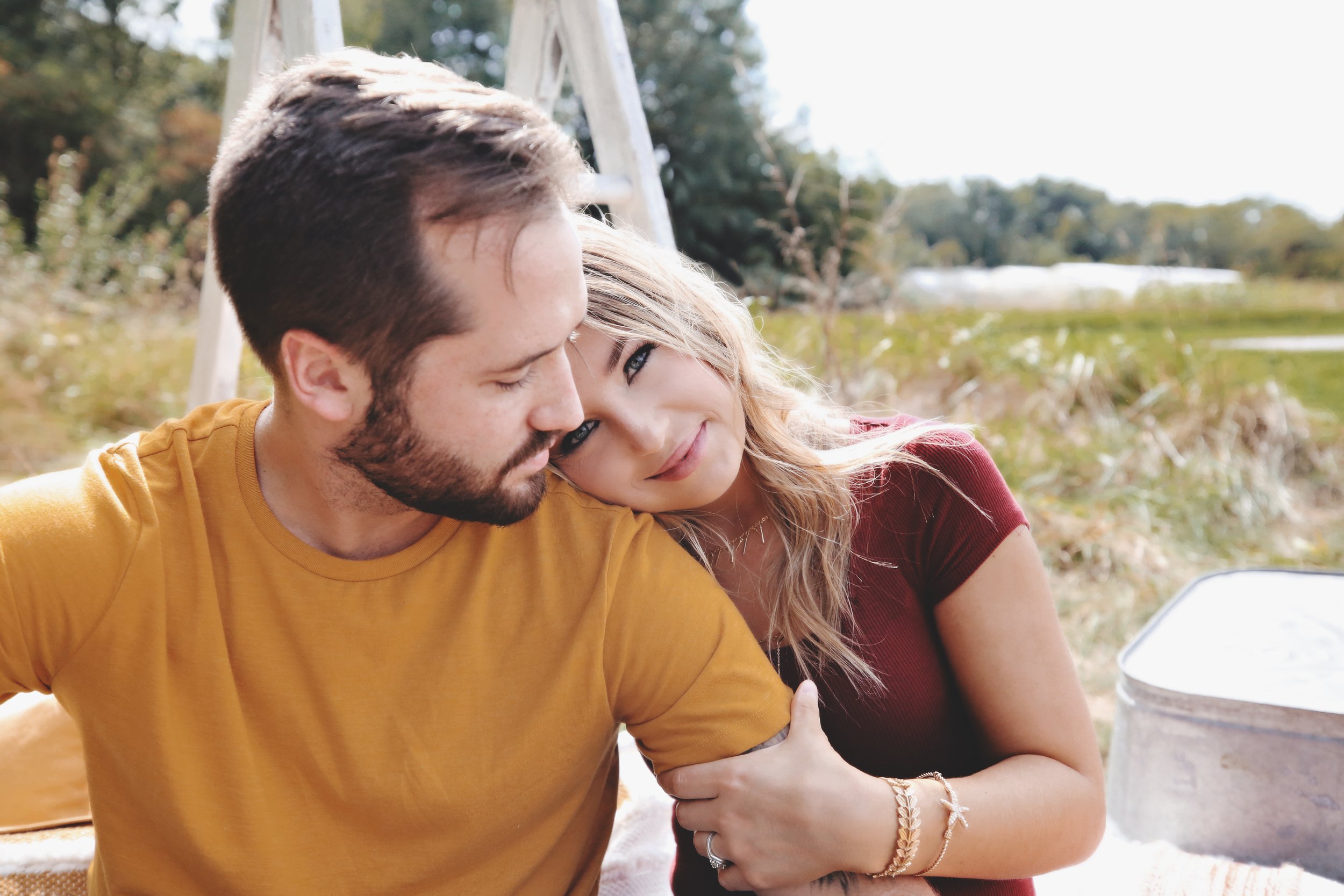 A happy couple sitting close together outdoors during daytime, with the woman resting her head on the man's shoulder and smiling, in a natural setting with trees and a field in the background.