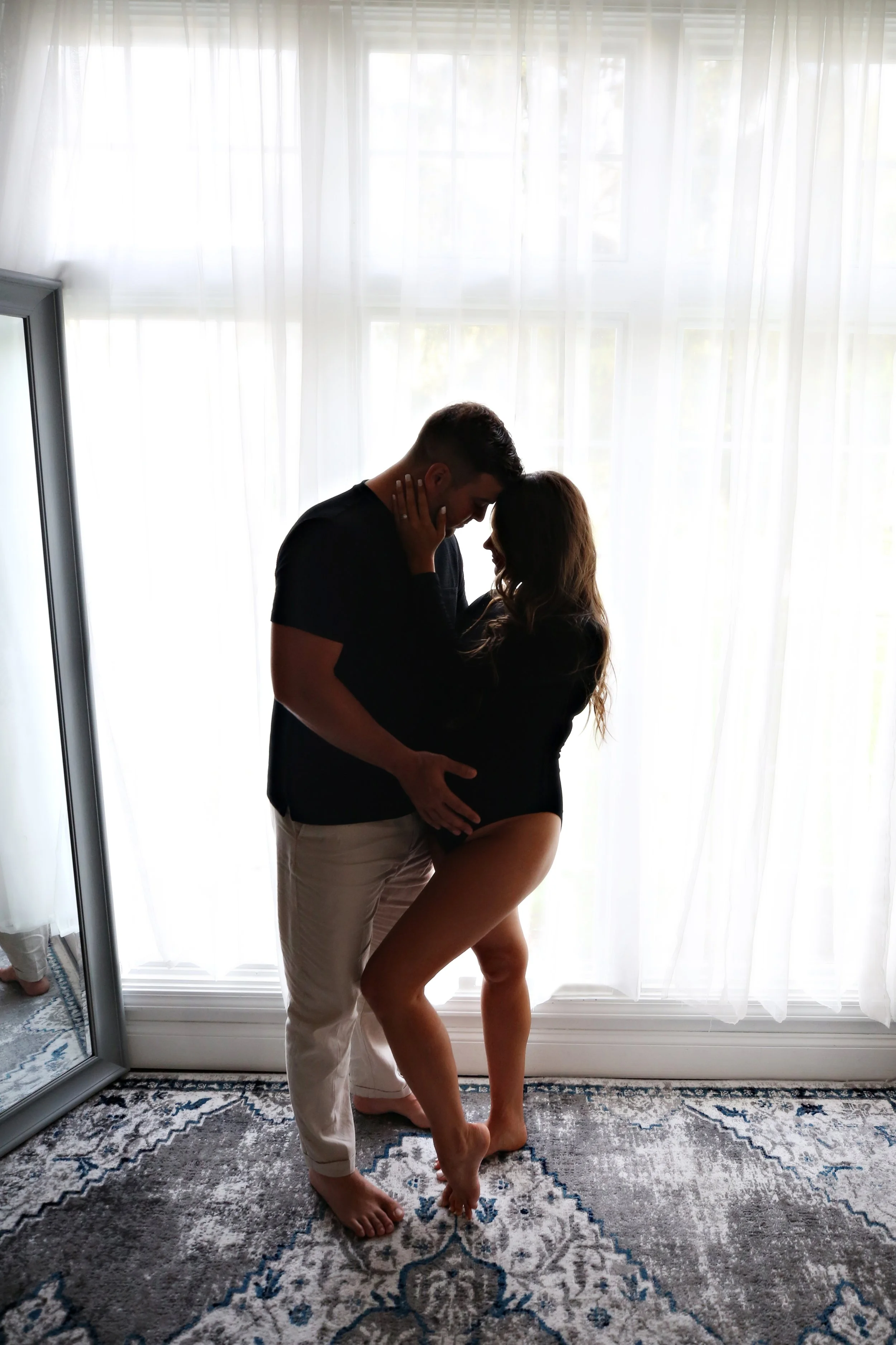 Silhouette of a romantic couple in an intimate moment, standing close together in front of bright sheer curtains, with the woman barefoot on a patterned rug.