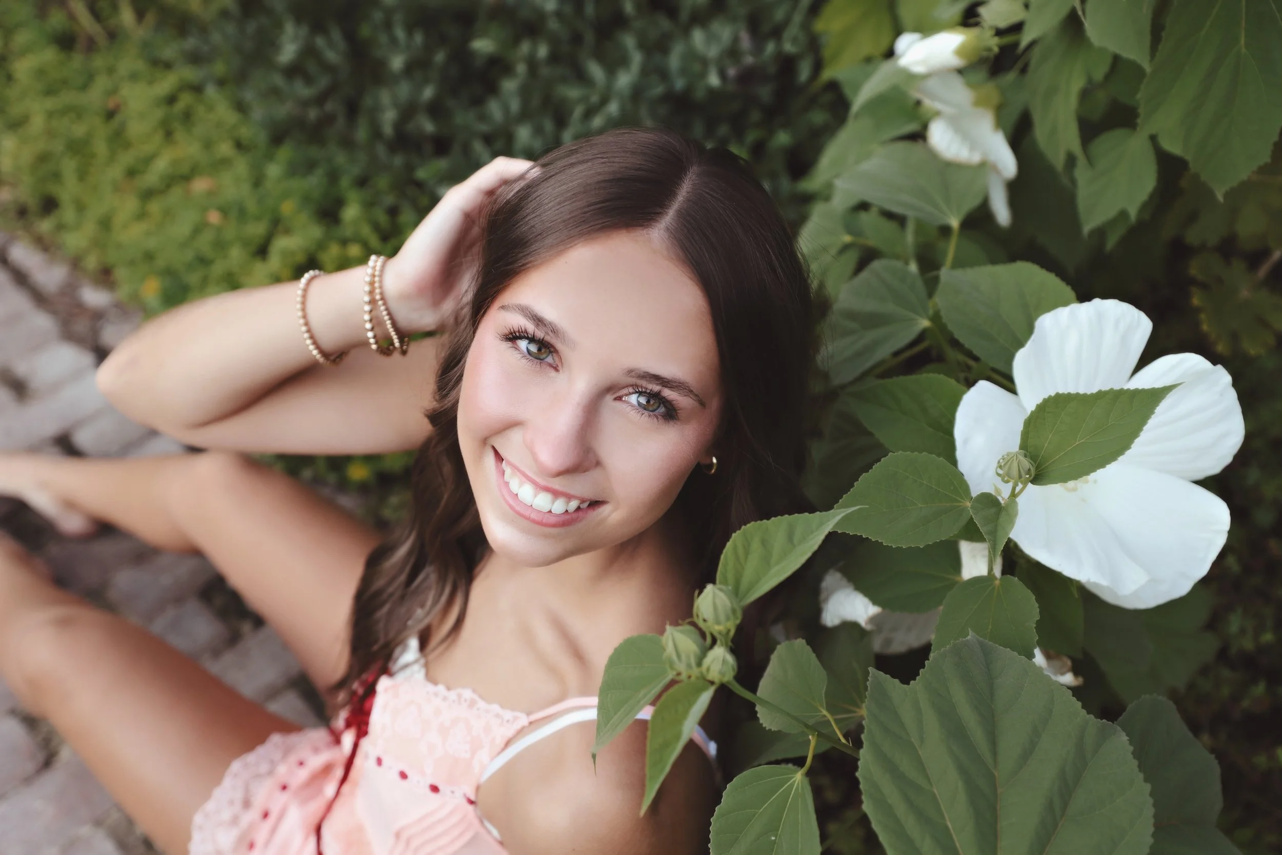 Young woman smiling while sitting on a cobblestone path next to white flowers and green leaves.