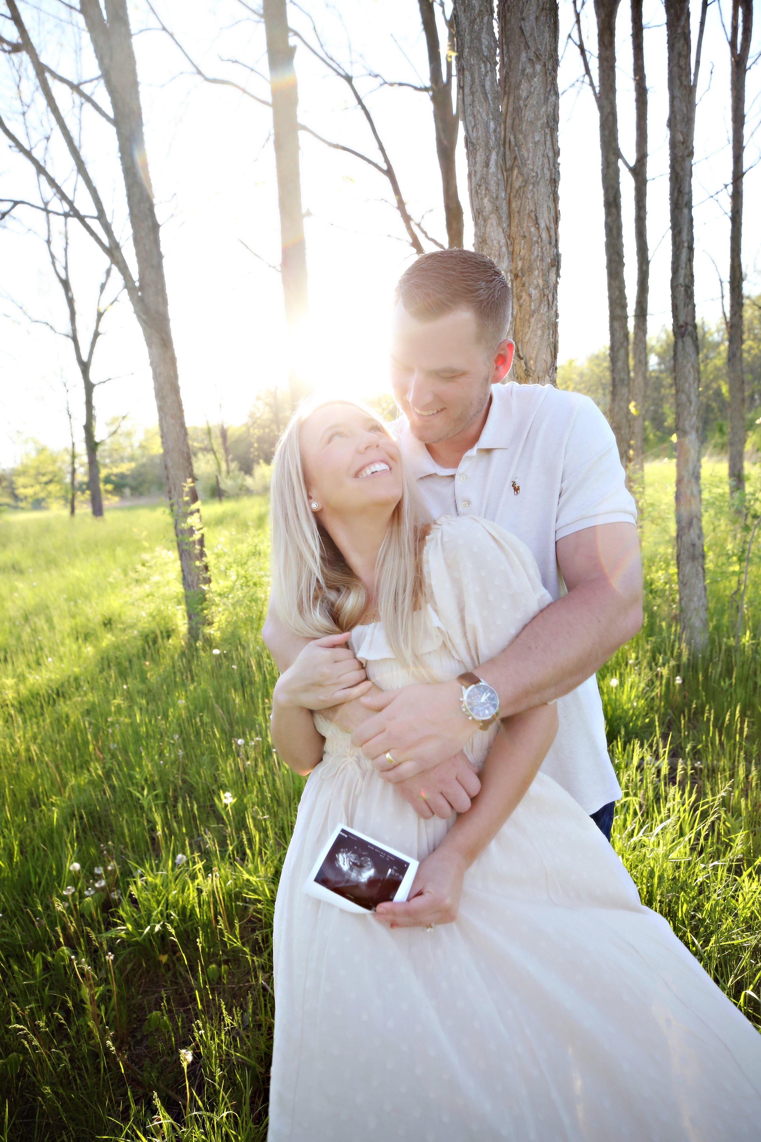 A couple in a sunlit forest with a woman holding an ultrasound image, smiling joyfully, as the man embraces her from behind.