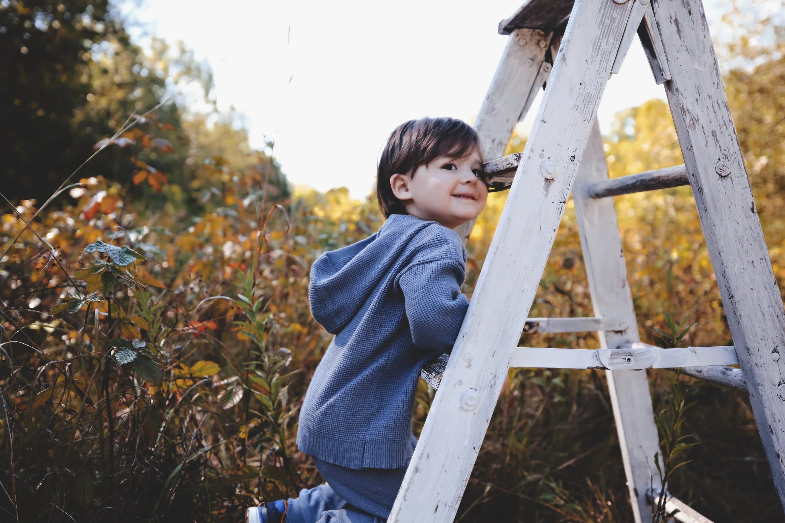 Young boy with dark hair climbing a white wooden ladder outdoors during autumn, surrounded by colorful fall foliage, smiling at the camera.