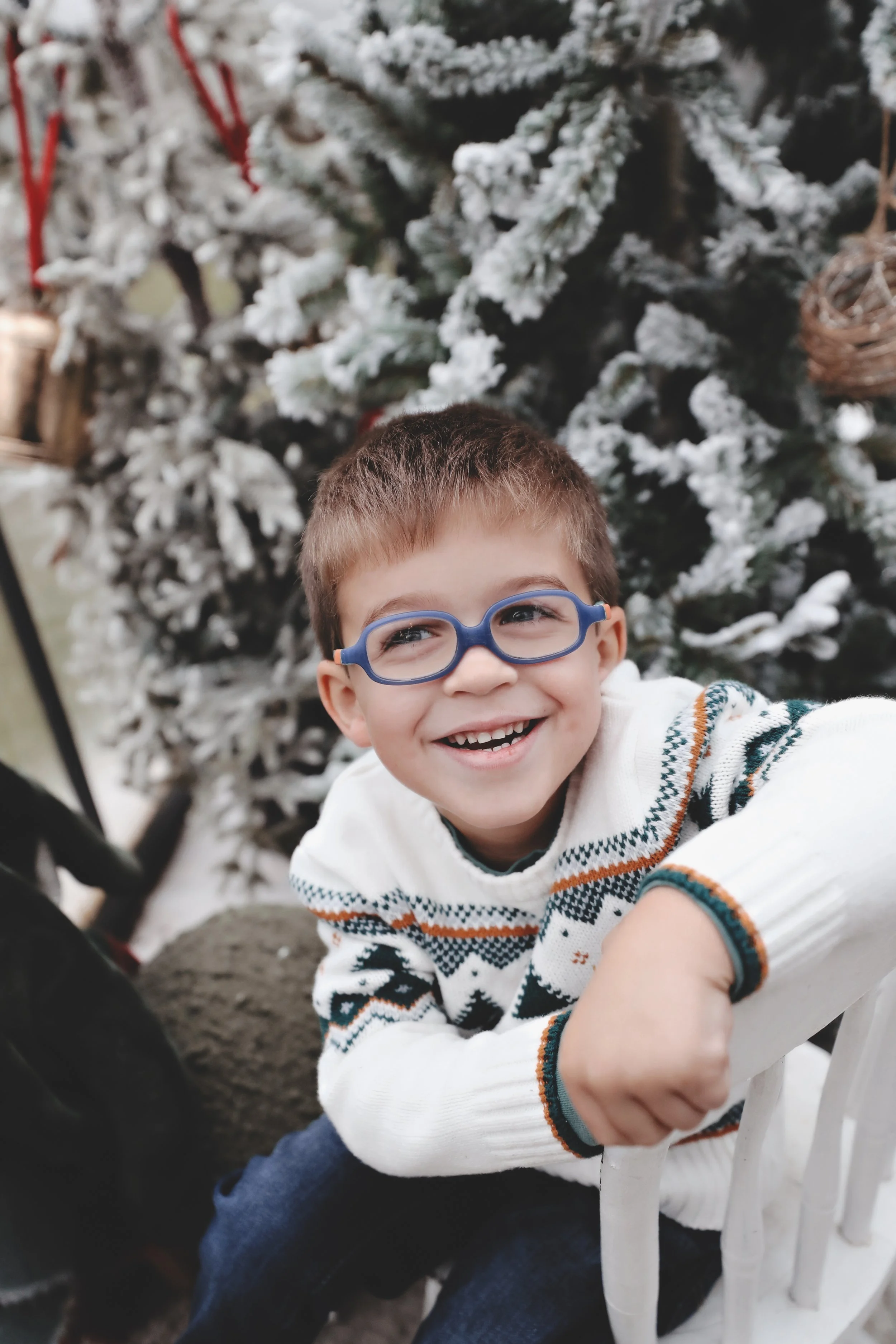 A young boy wearing glasses and a holiday sweater, smiling and leaning on a white chair in front of a snow-covered Christmas tree.