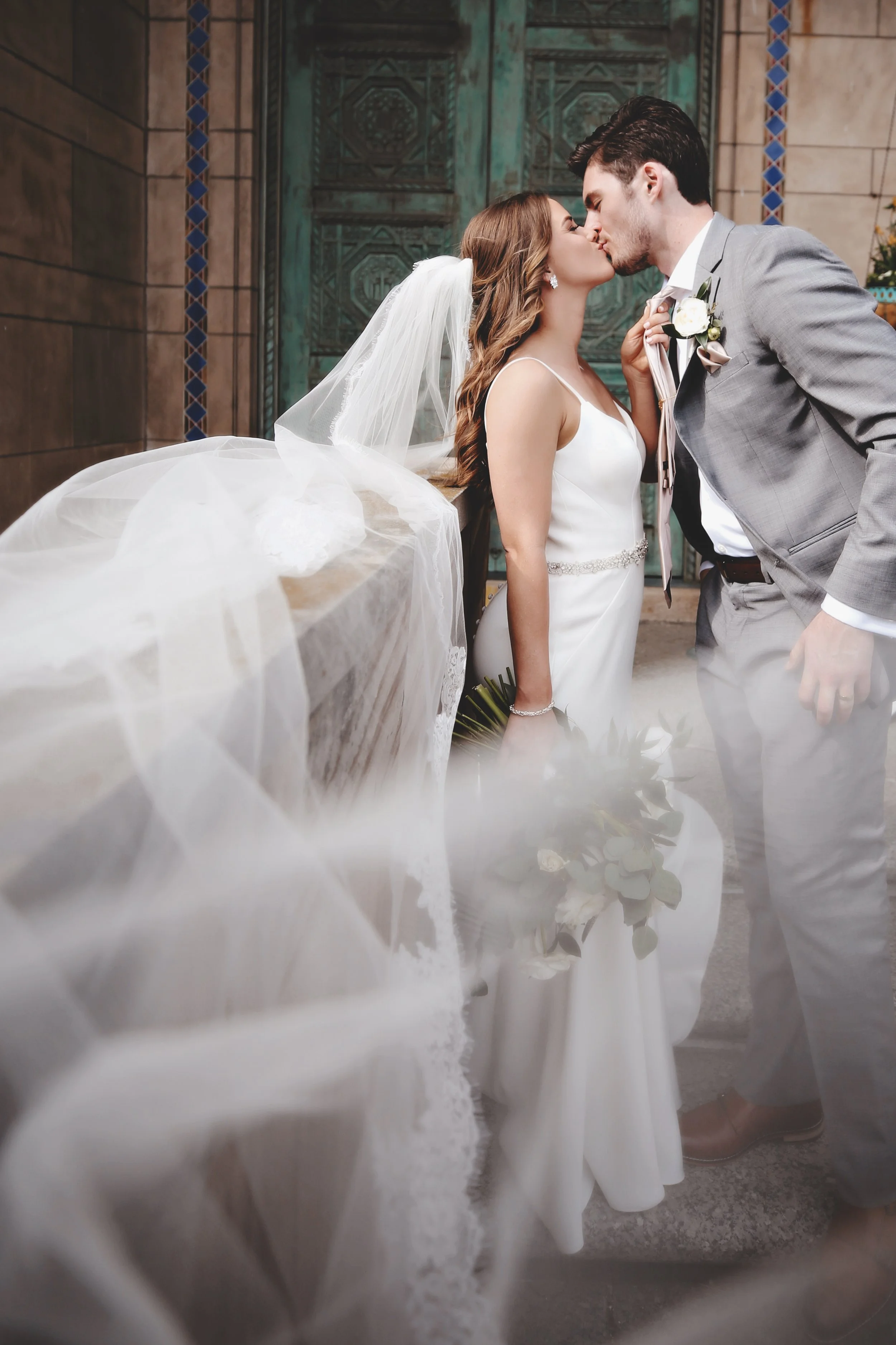 A bride and groom kiss at their wedding, with the bride wearing a white gown and veil, and the groom in a gray suit with a boutonniere, against a decorative backdrop.