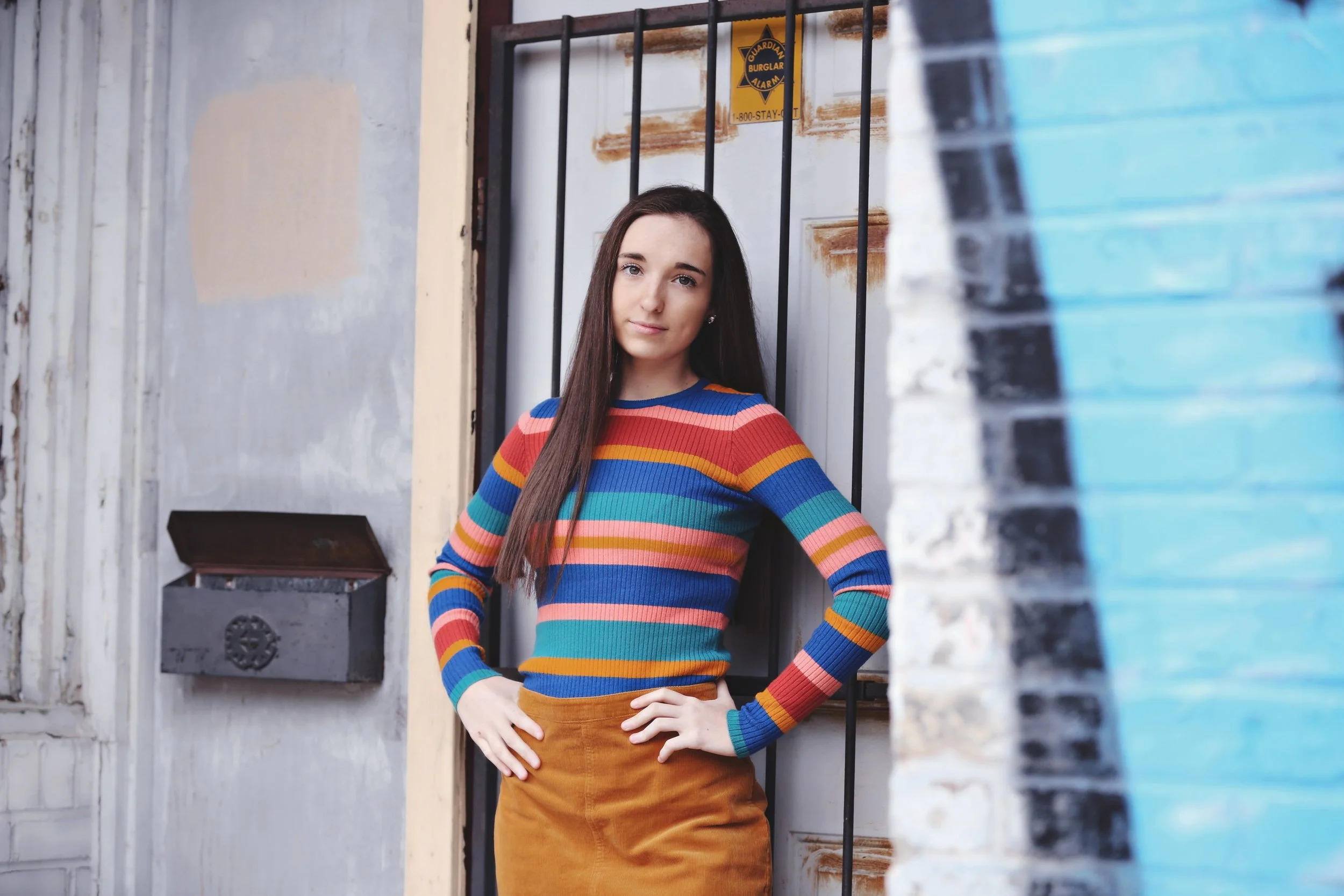 A young woman wearing a colorful striped sweater and brown skirt standing against a metal gate and wall in an urban setting.
