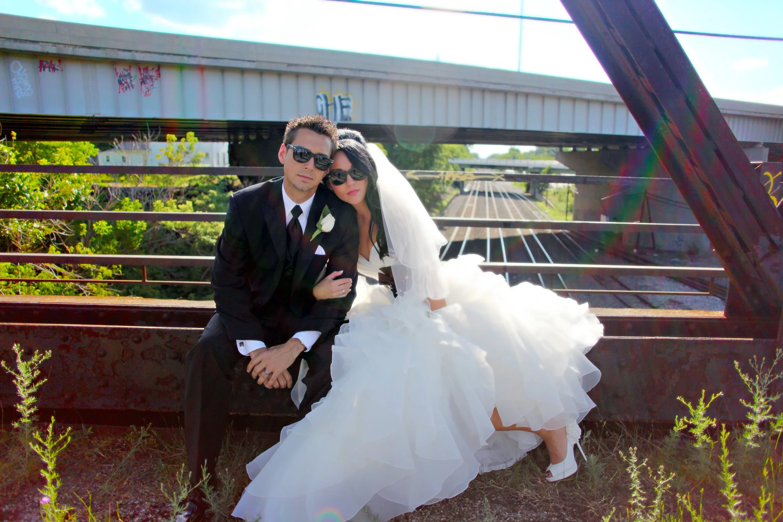 A bride and groom sit on a rusted railway bridge with graffiti, sunlight shining, greenery in the background, both wearing sunglasses, the bride in a white wedding gown and veil, the groom in a black tuxedo with a white flower boutonniere.