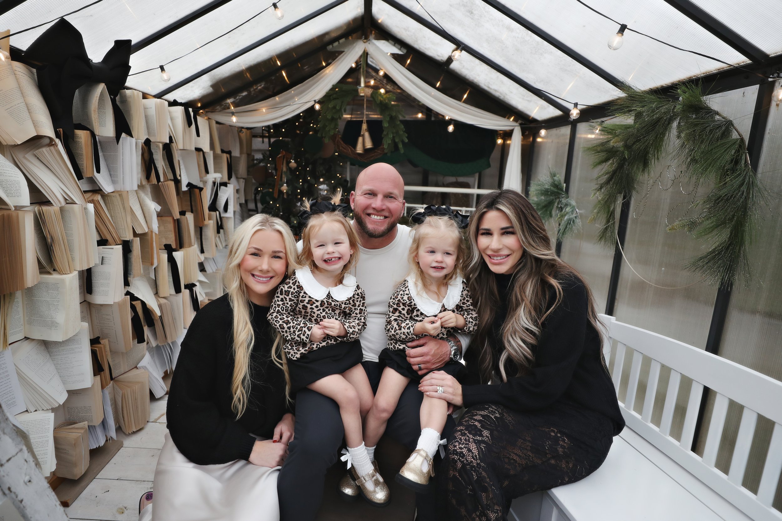 A family of five sitting inside a decorated greenhouse with books on the walls, Christmas trees, and string lights, all smiling at the camera.