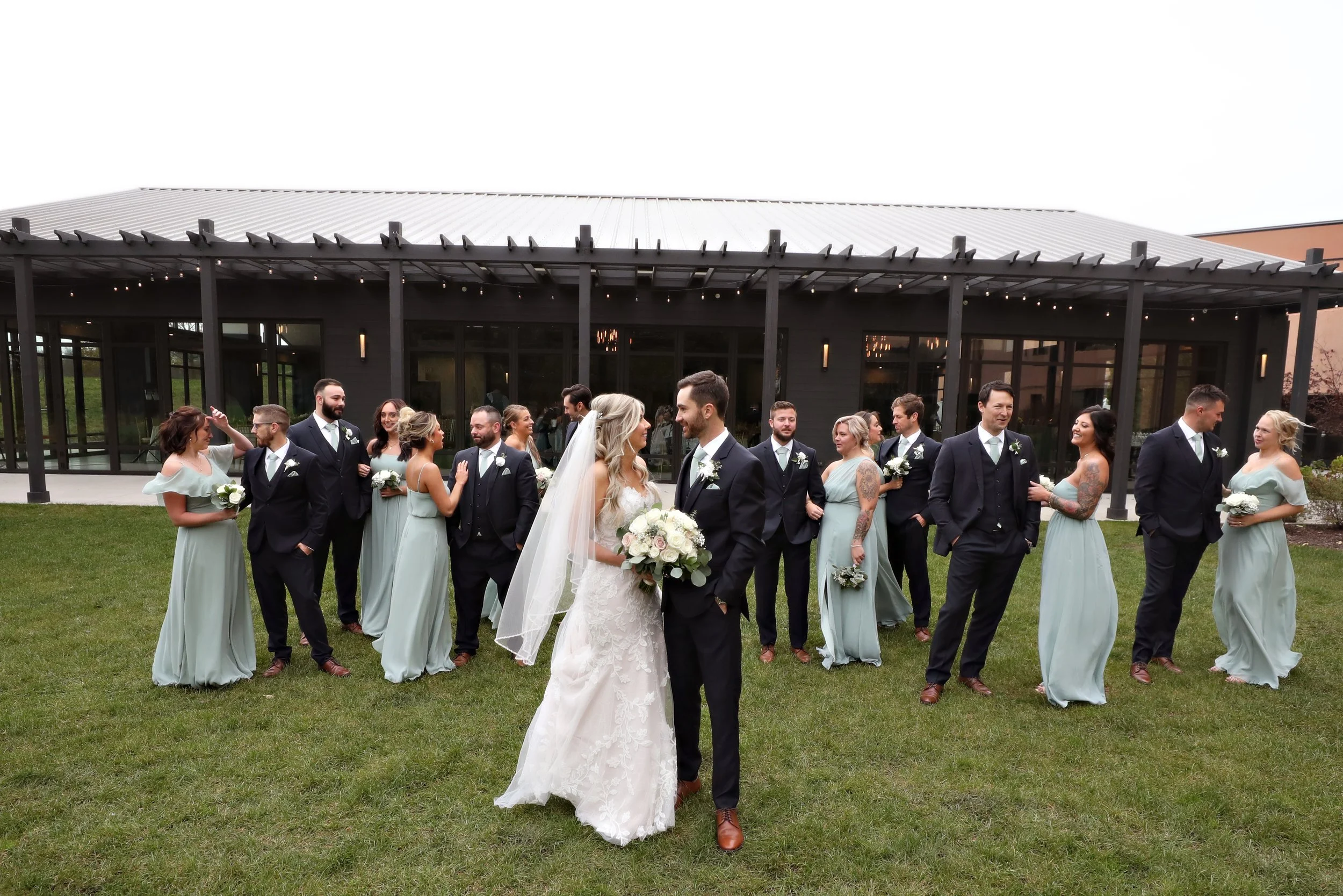 Wedding party outdoors with bride and groom in center, surrounded by bridesmaids and groomsmen on a grassy area in front of a modern building.