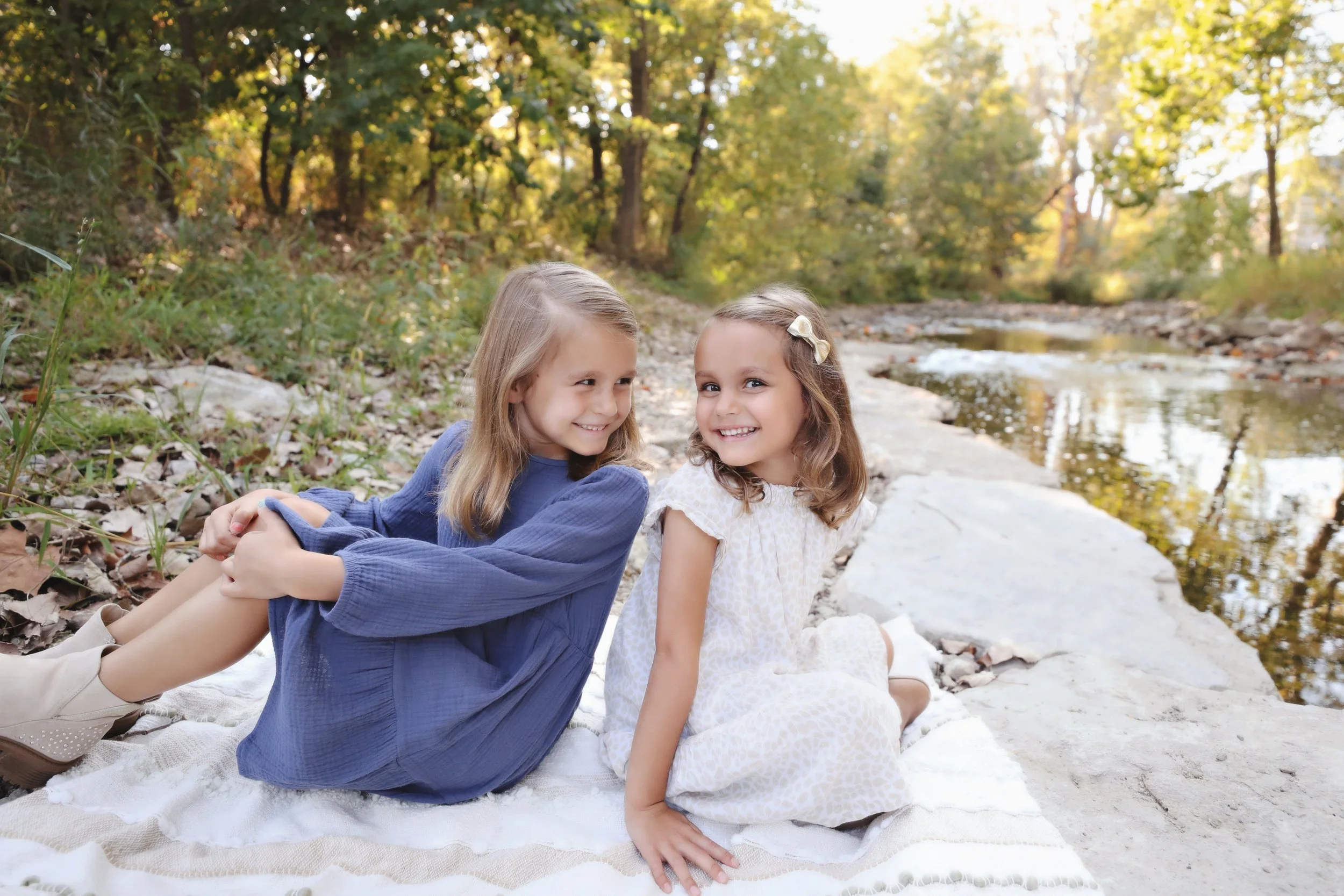 Two young girls sitting on a blanket near a creek in a forest, smiling at each other.