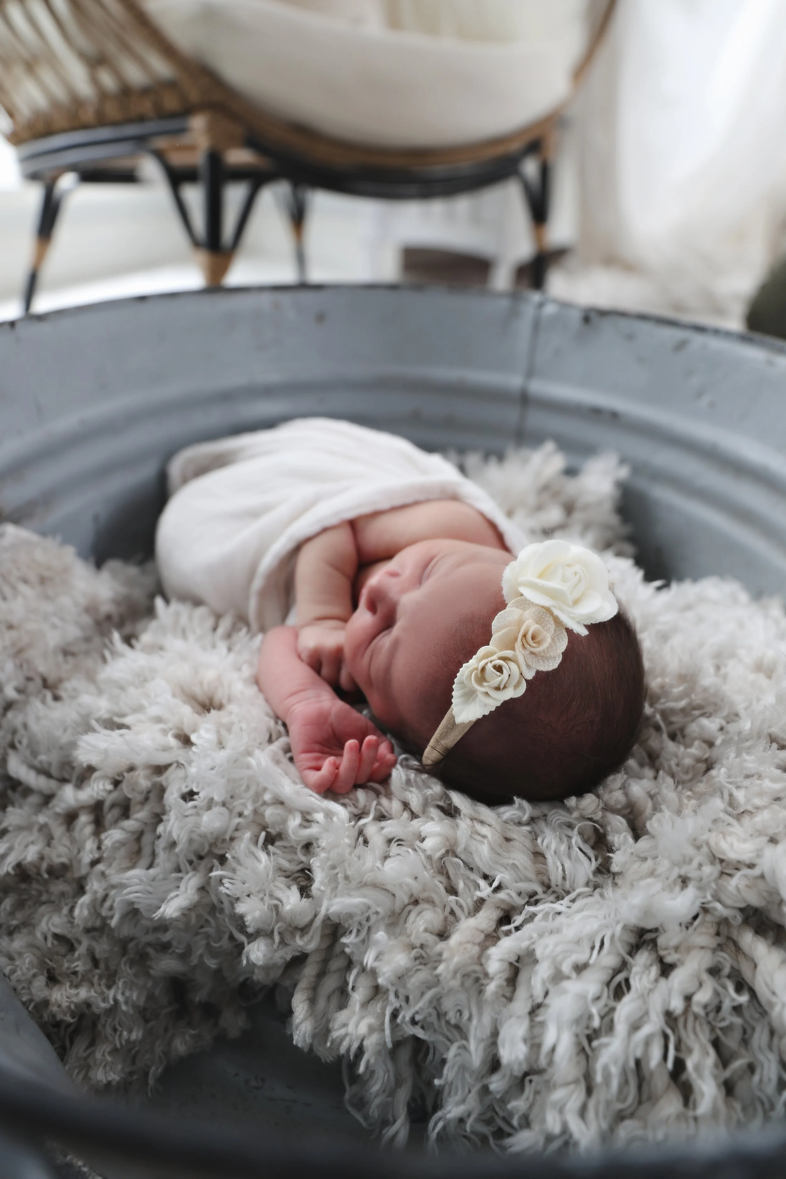 Newborn baby sleeping on a soft, fluffy blanket in a metal tub, wearing a headband with white flowers.