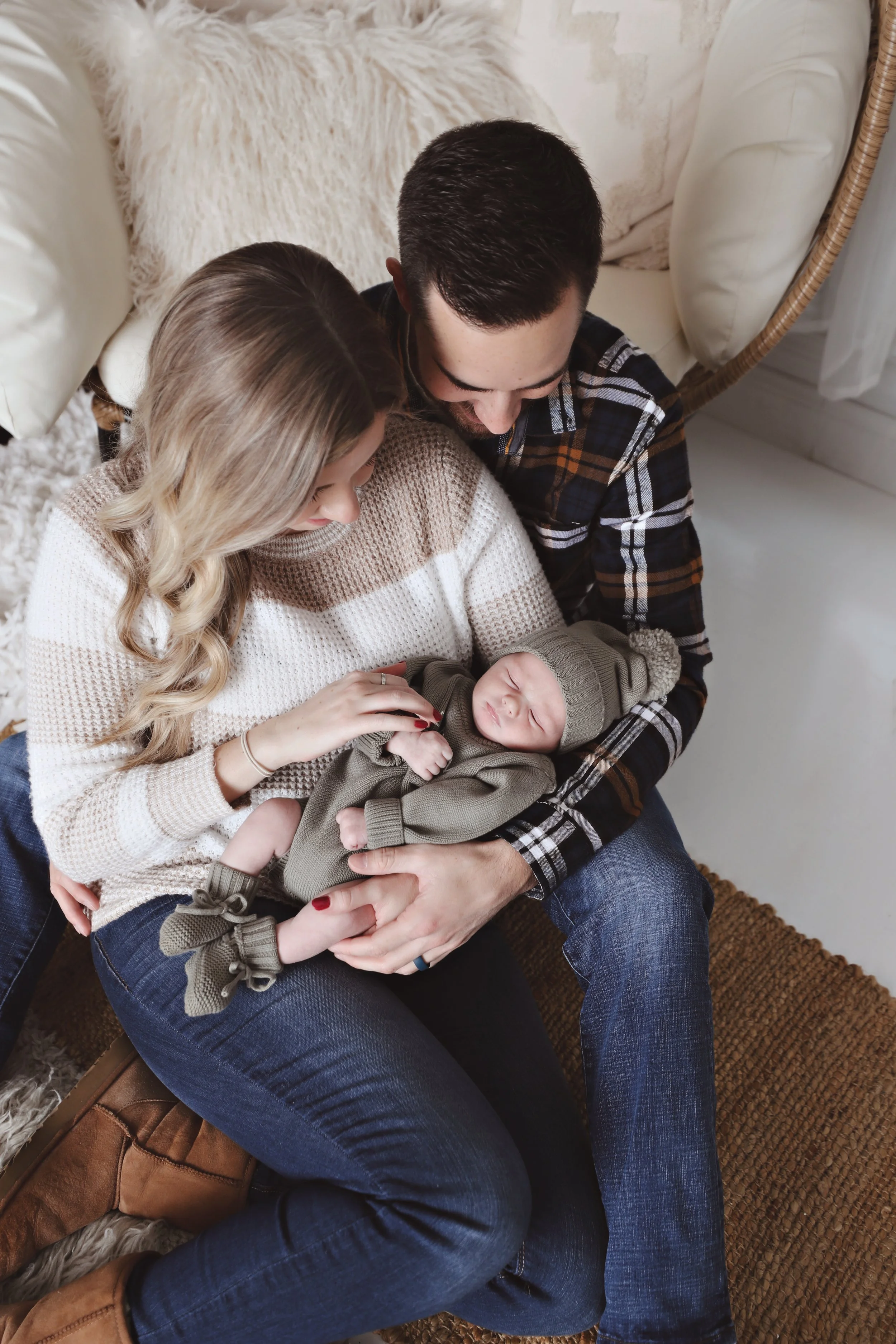 A family of three sitting on a rug, holding a sleeping baby. The mother has long blonde hair and wears a beige sweater, the father has short dark hair and wears a plaid shirt, and the baby is dressed in gray with a hat.