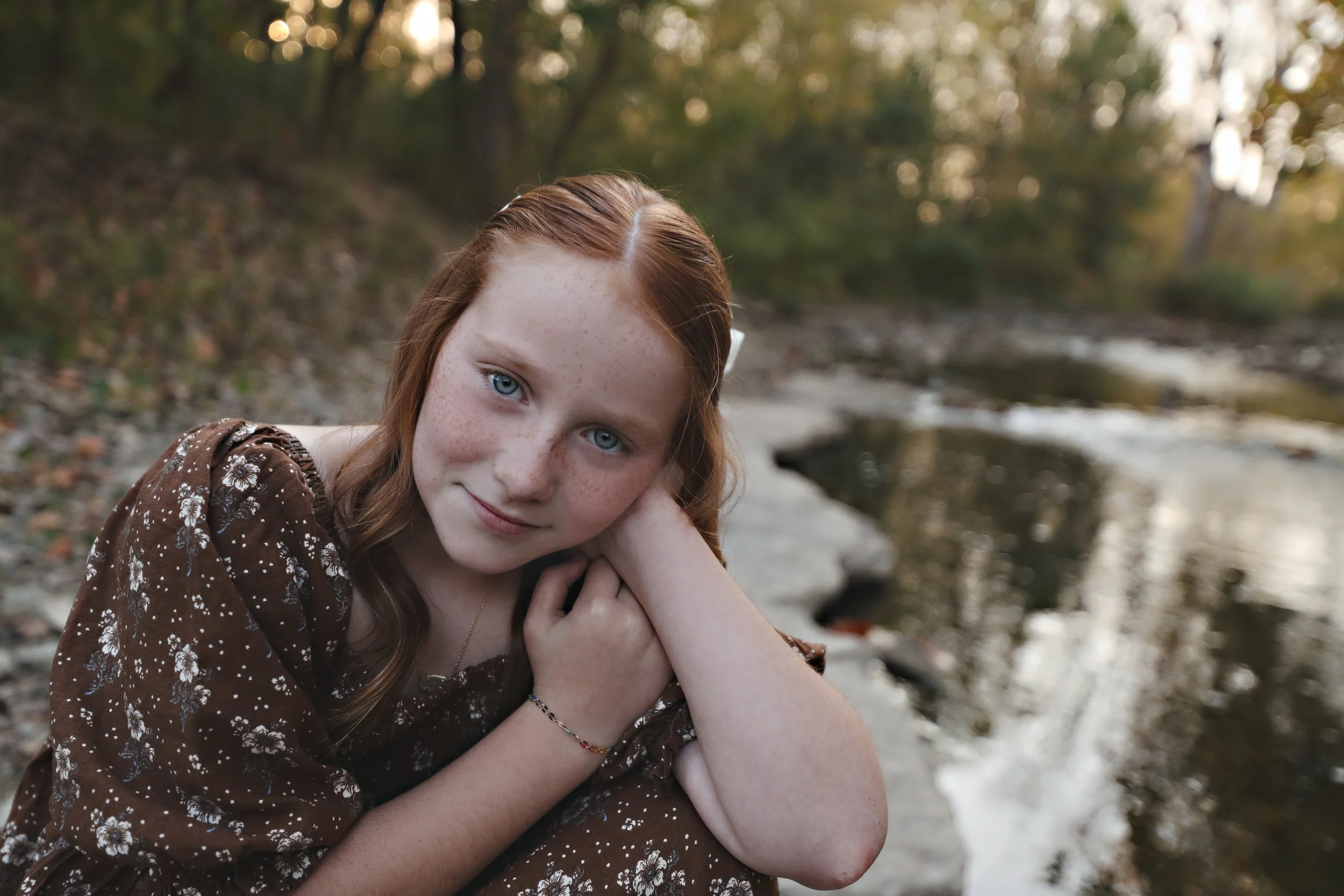 A young woman with red hair and blue eyes sitting near a riverbank during autumn, resting her head on her hand and smiling gently at the camera.