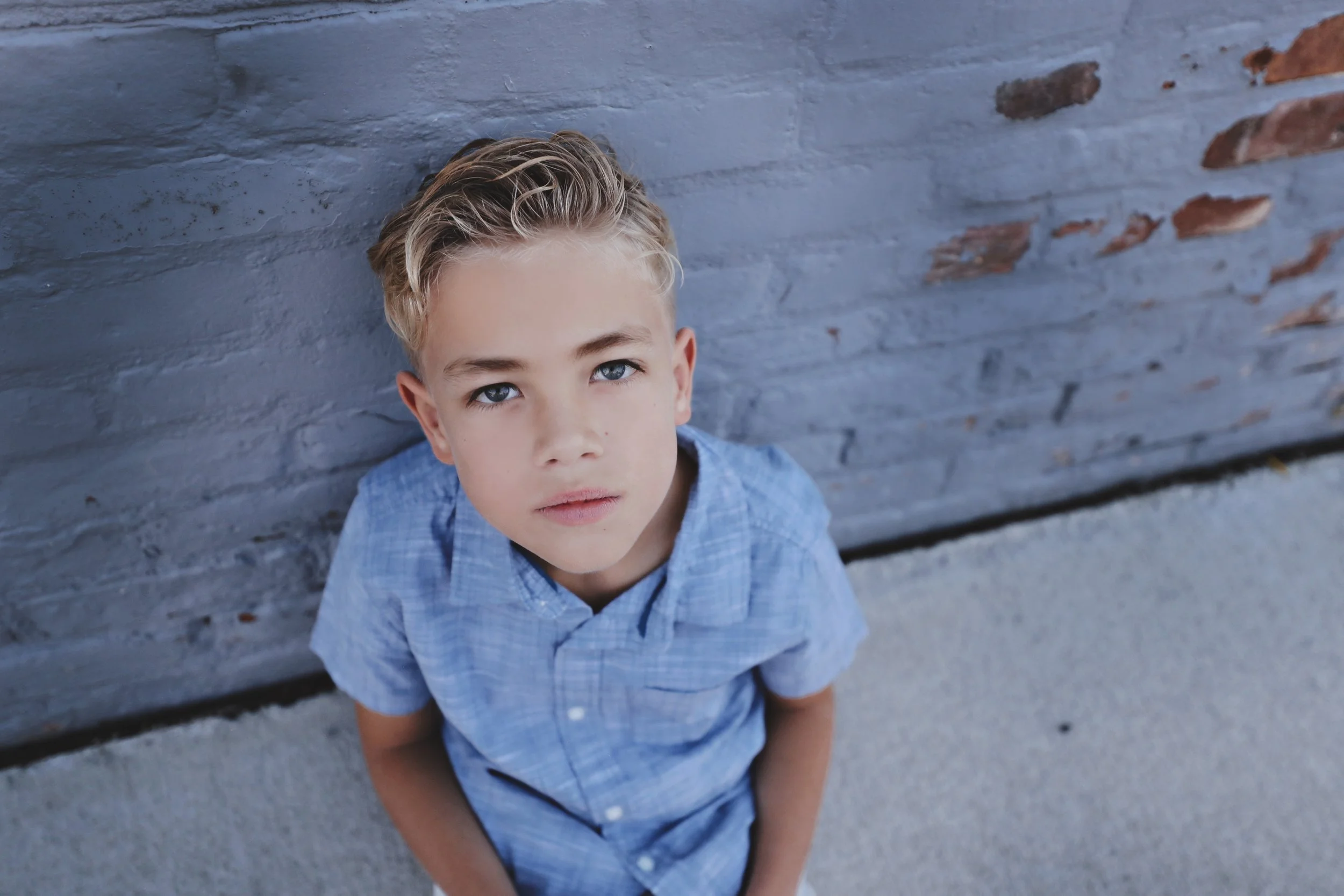 A young boy with blonde hair and blue eyes, wearing a blue shirt, sitting against a gray brick wall, looking up at the camera with a serious expression.