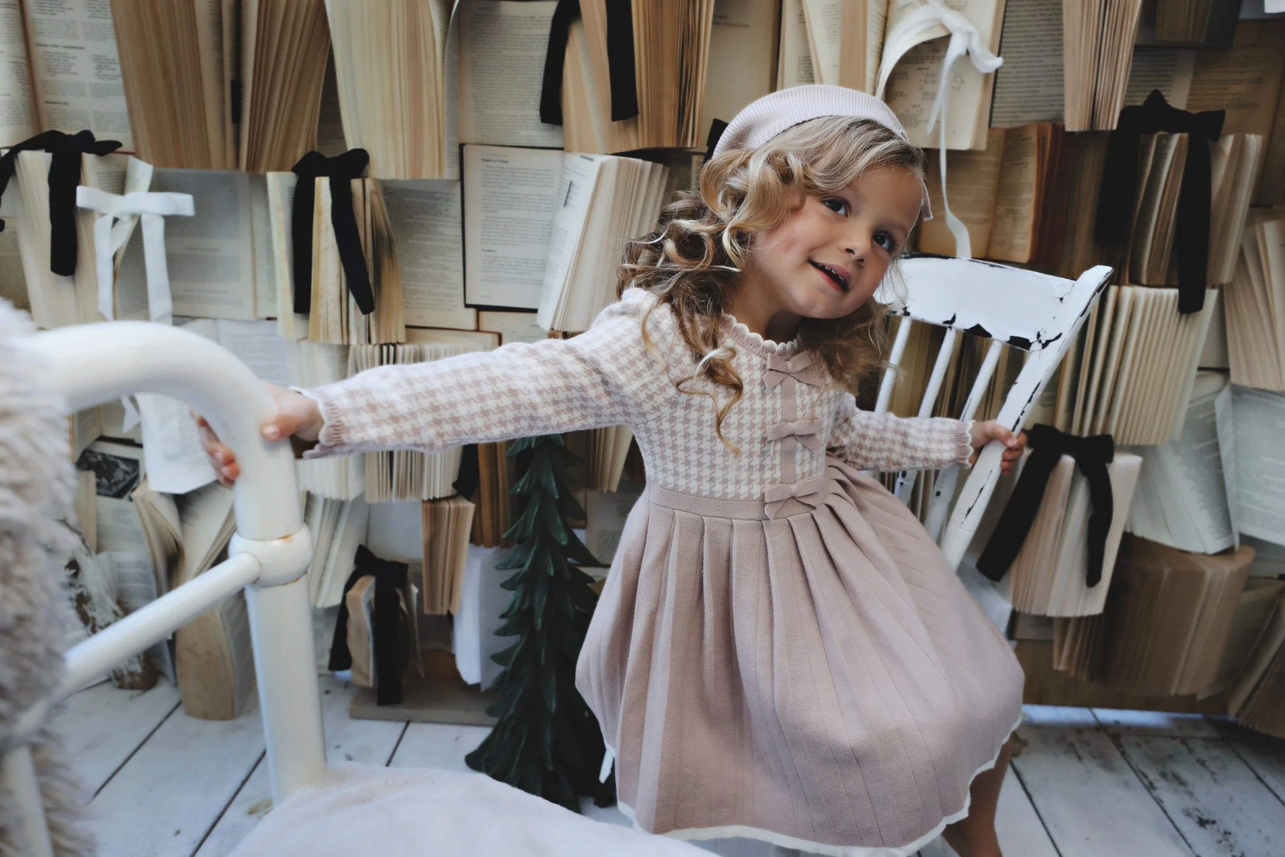 A young girl with curly hair wearing a pink dress and a white bonnet sitting on a white chair in front of a background filled with open books with black ribbons tied around some. She is smiling and holding onto the armrests of the chair.