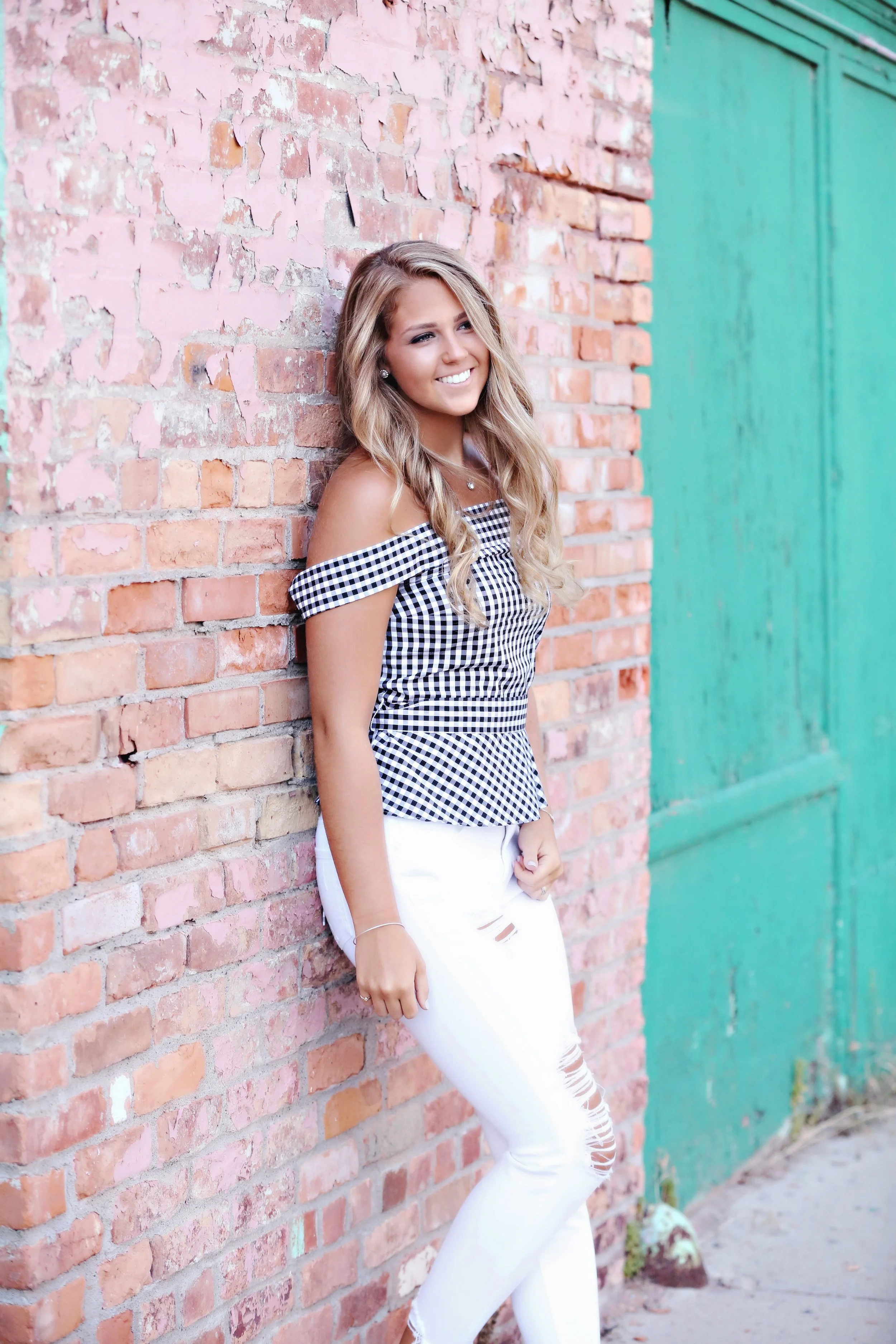 A young woman with long wavy blonde hair, smiling, standing against a pink brick wall with a teal door next to her.