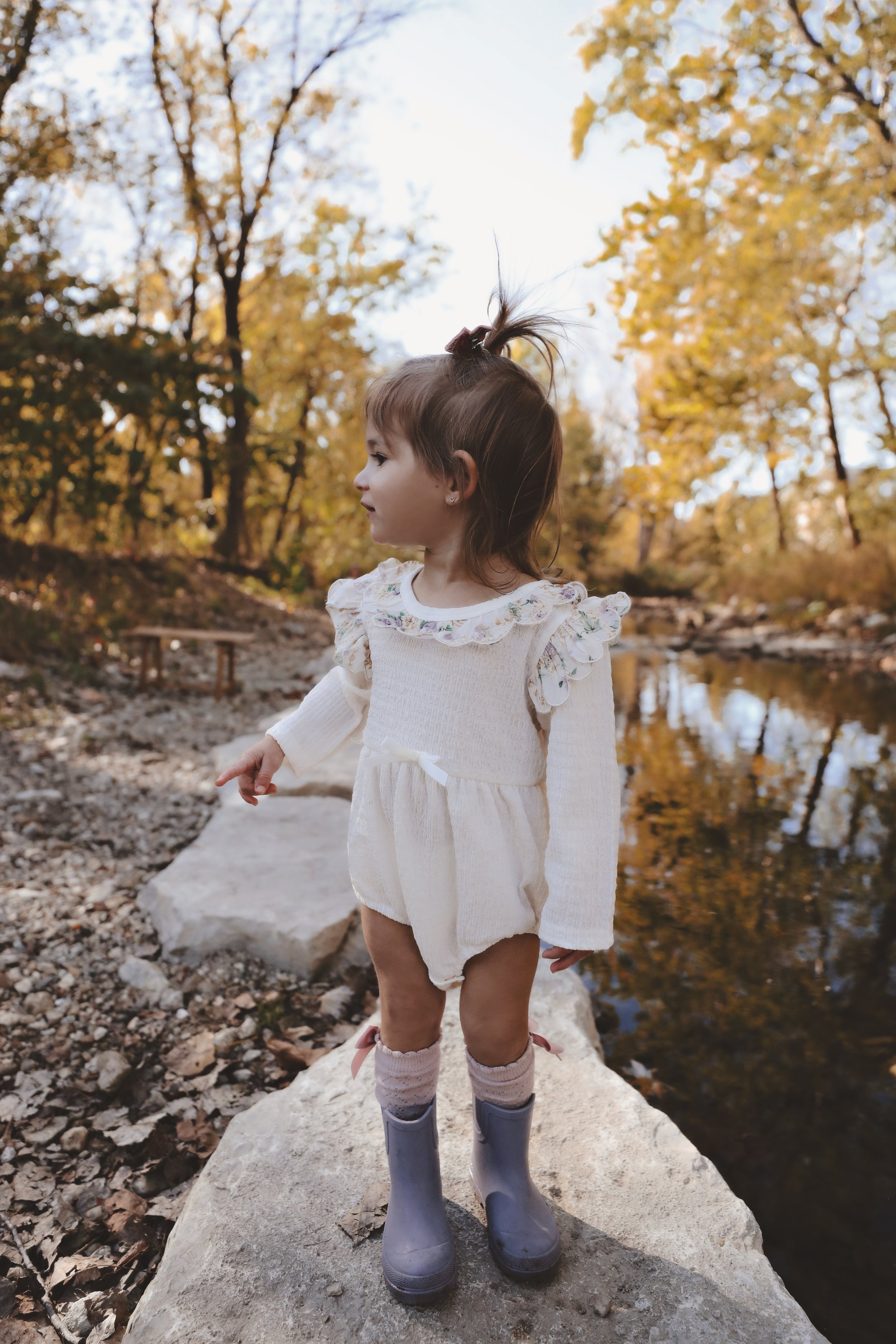A young girl with brown hair standing on a rock by a creek in a forest with fall-colored leaves.