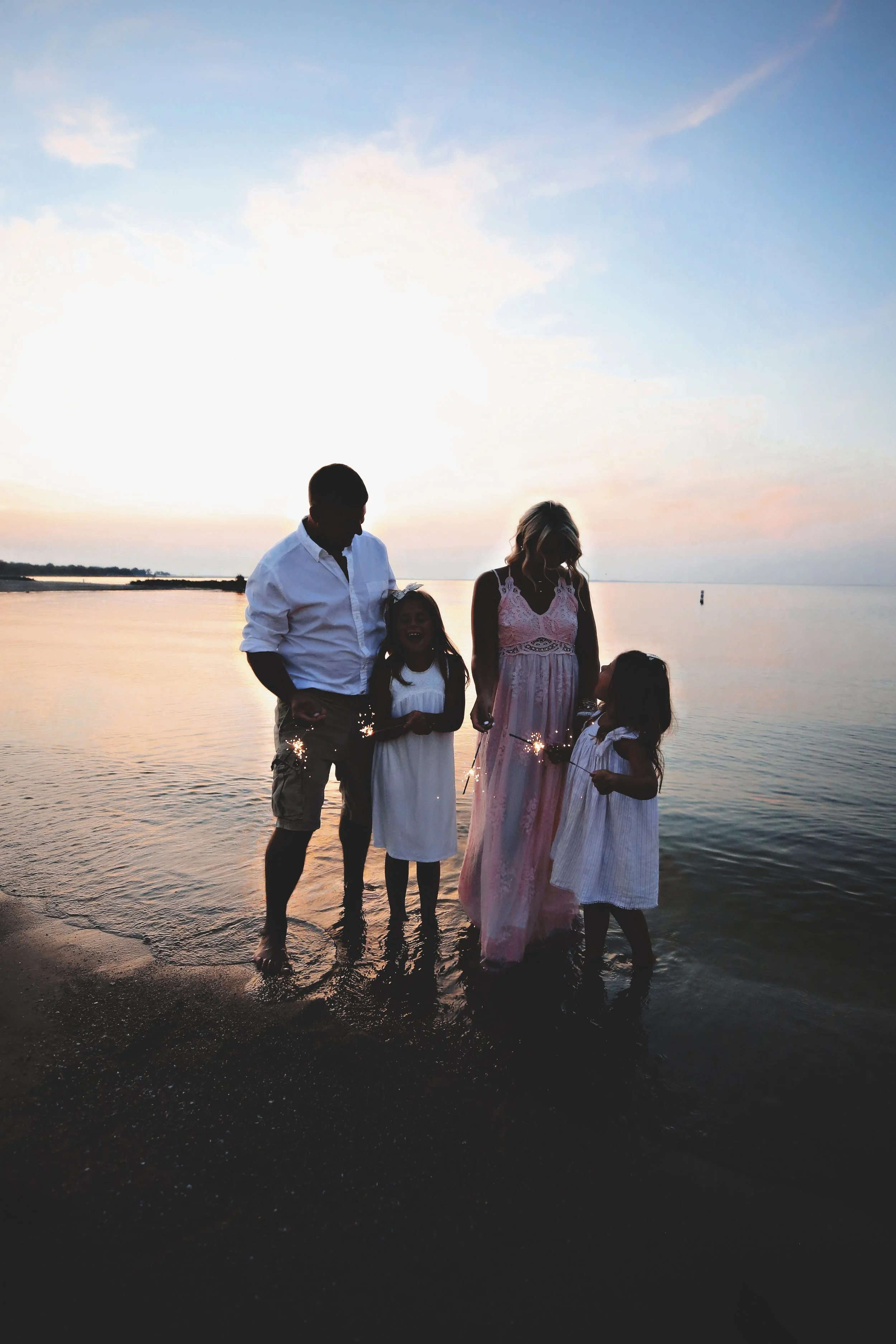 Family standing at the edge of the water during sunset, holding sparklers.