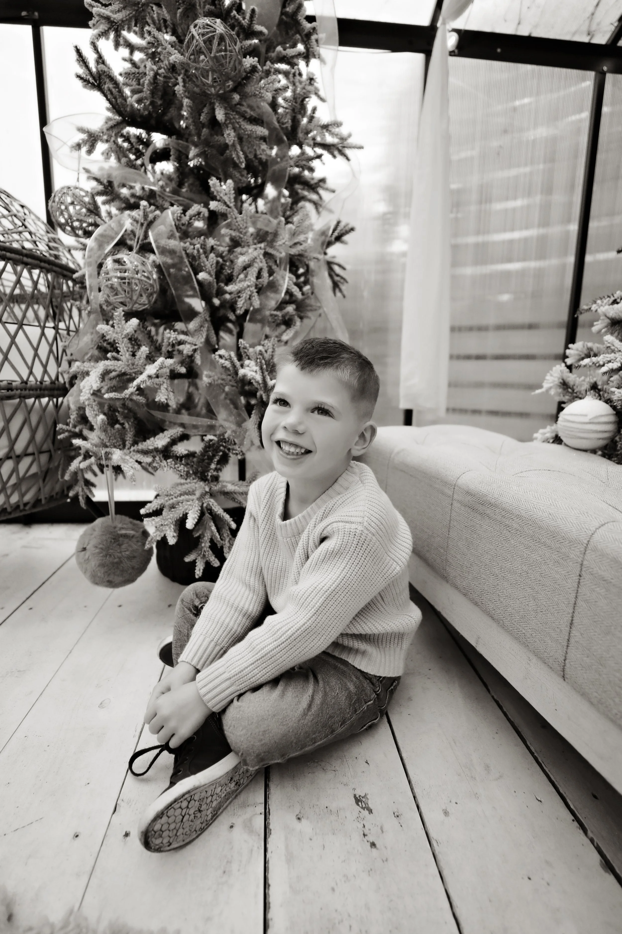 A young boy sitting on the wooden floor, smiling, near a decorated Christmas tree with ornaments, in black and white.