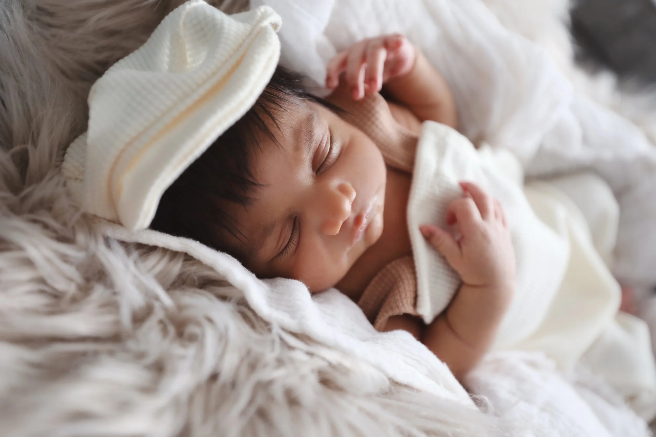 A sleeping baby wearing a white hat with yellow stripes, lying on a fluffy beige blanket.