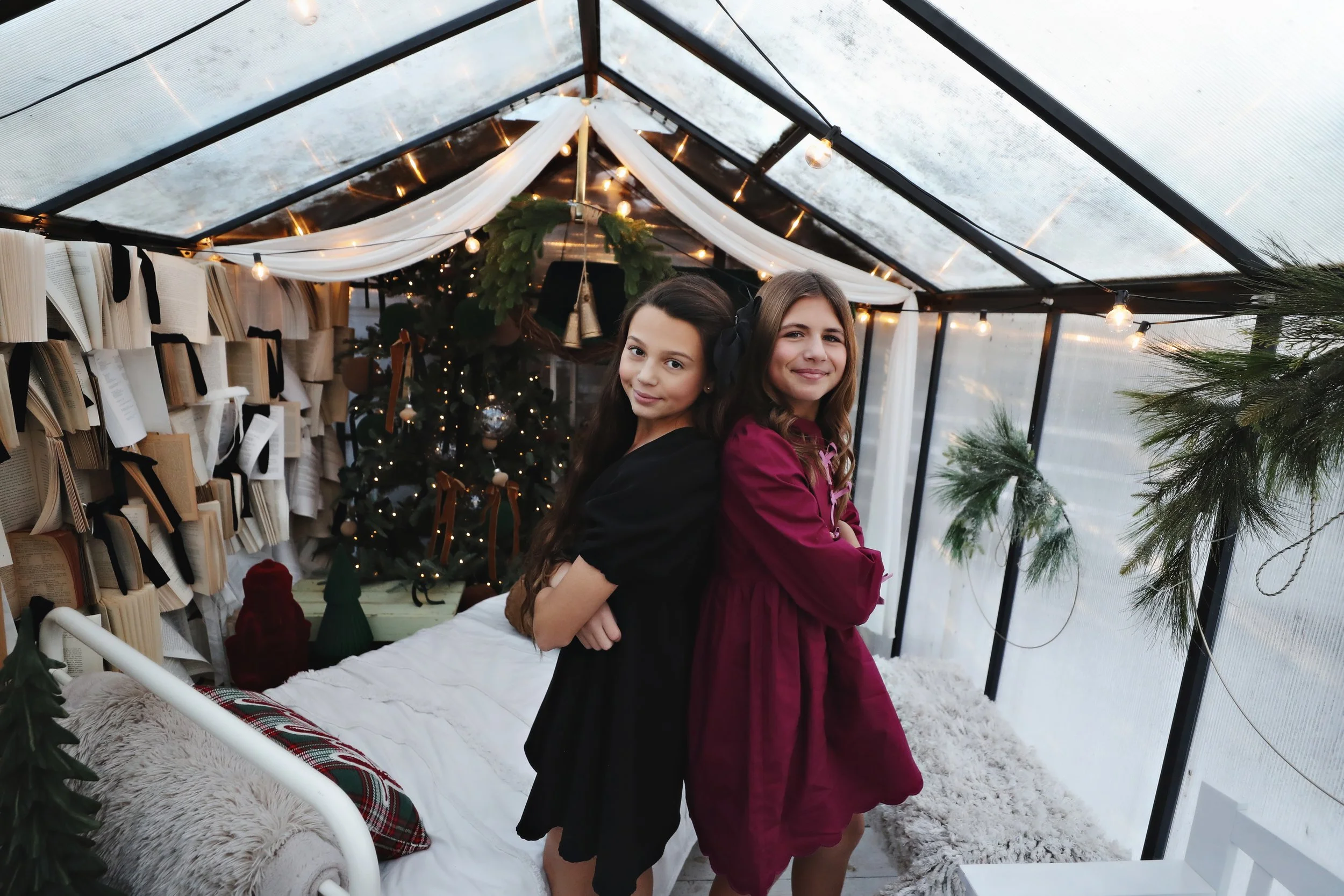 Two young girls standing back-to-back inside a decorated glass greenhouse with Christmas lights, a Christmas tree, and books on the walls.