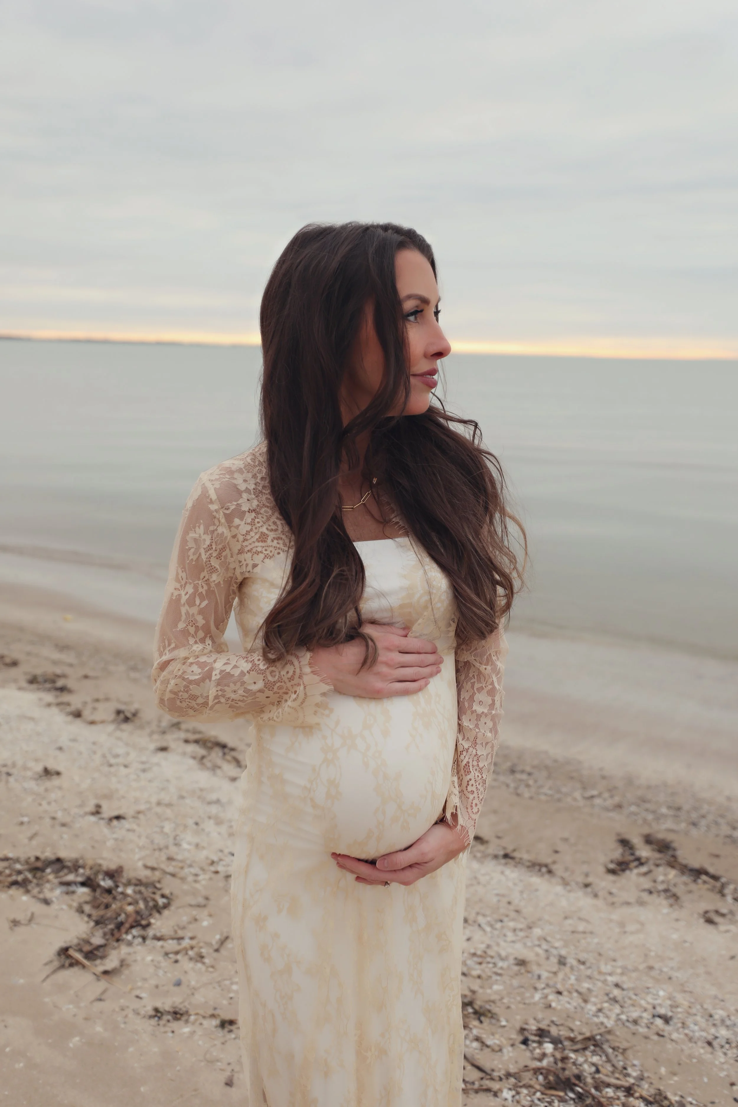 A pregnant woman with long dark hair in a lace dress standing on the beach, holding her belly with one hand, looking to the side, with calm water and a cloudy sky in the background.