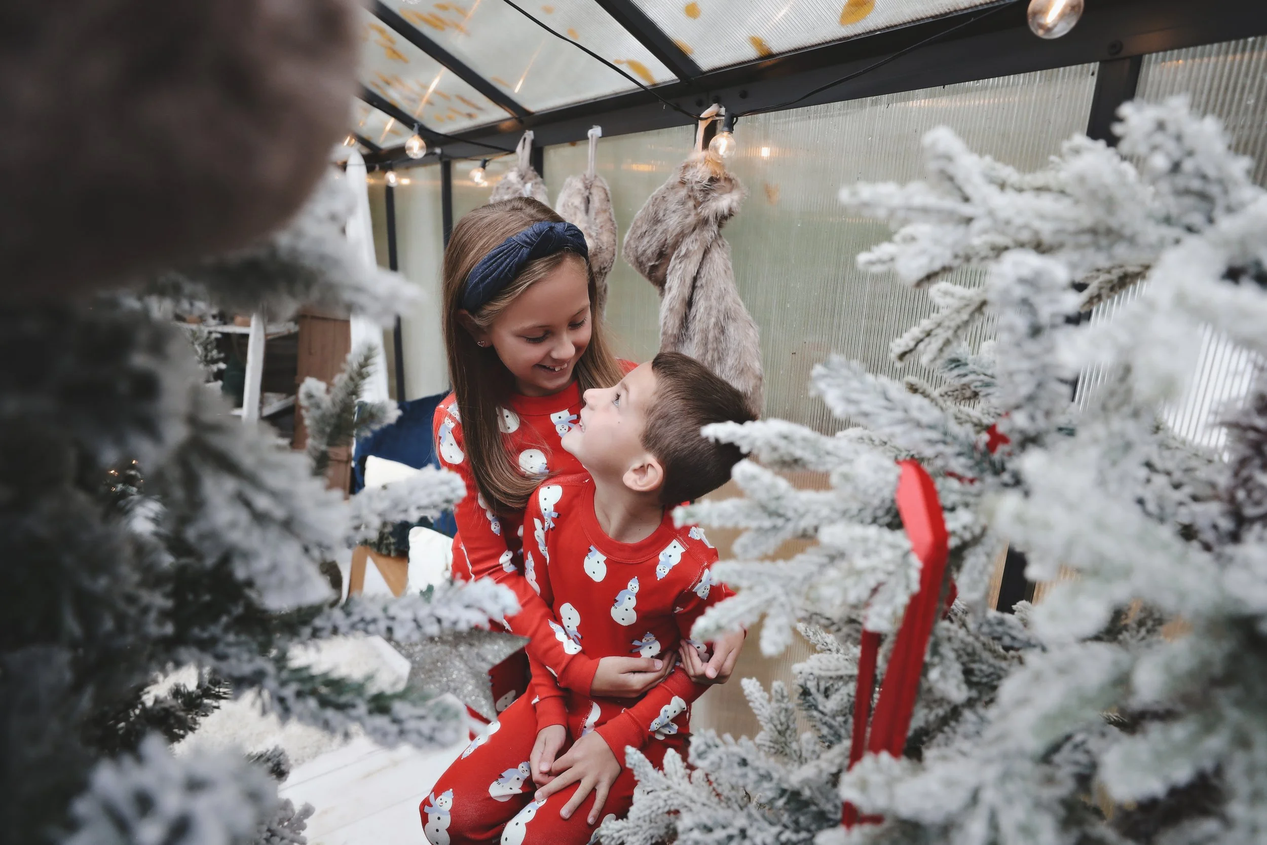 Two children in festive pajamas sharing a joyful moment amid snow-dusted Christmas trees inside a decorated greenhouse. The girl with a blue headband has her arms around the boy, and both are smiling at each other. Faux fur animal pelts hang in the b