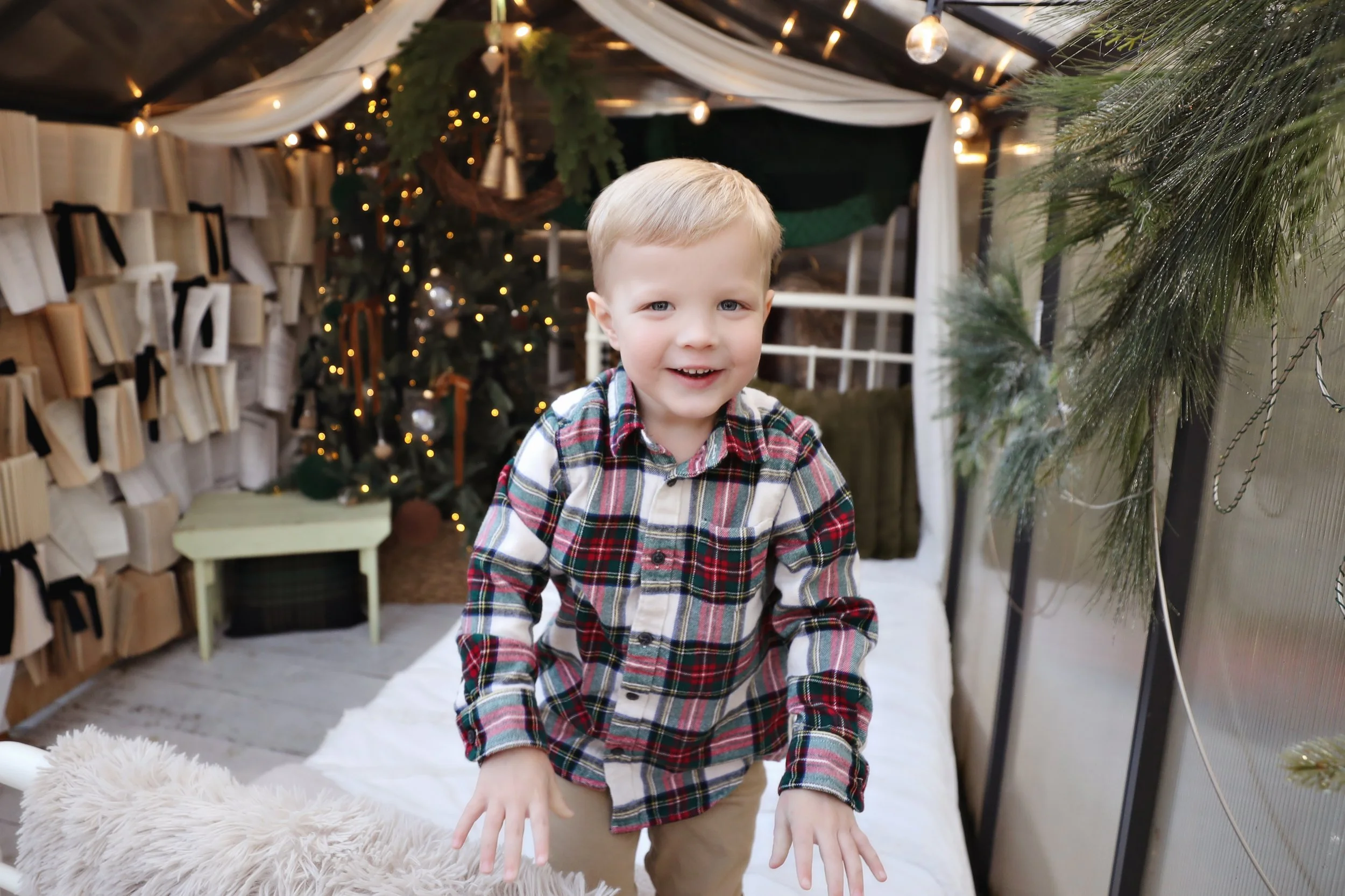 A young boy in a plaid shirt smiling and looking at the camera inside a festive decorated area with Christmas lights, a Christmas tree, and holiday decorations.