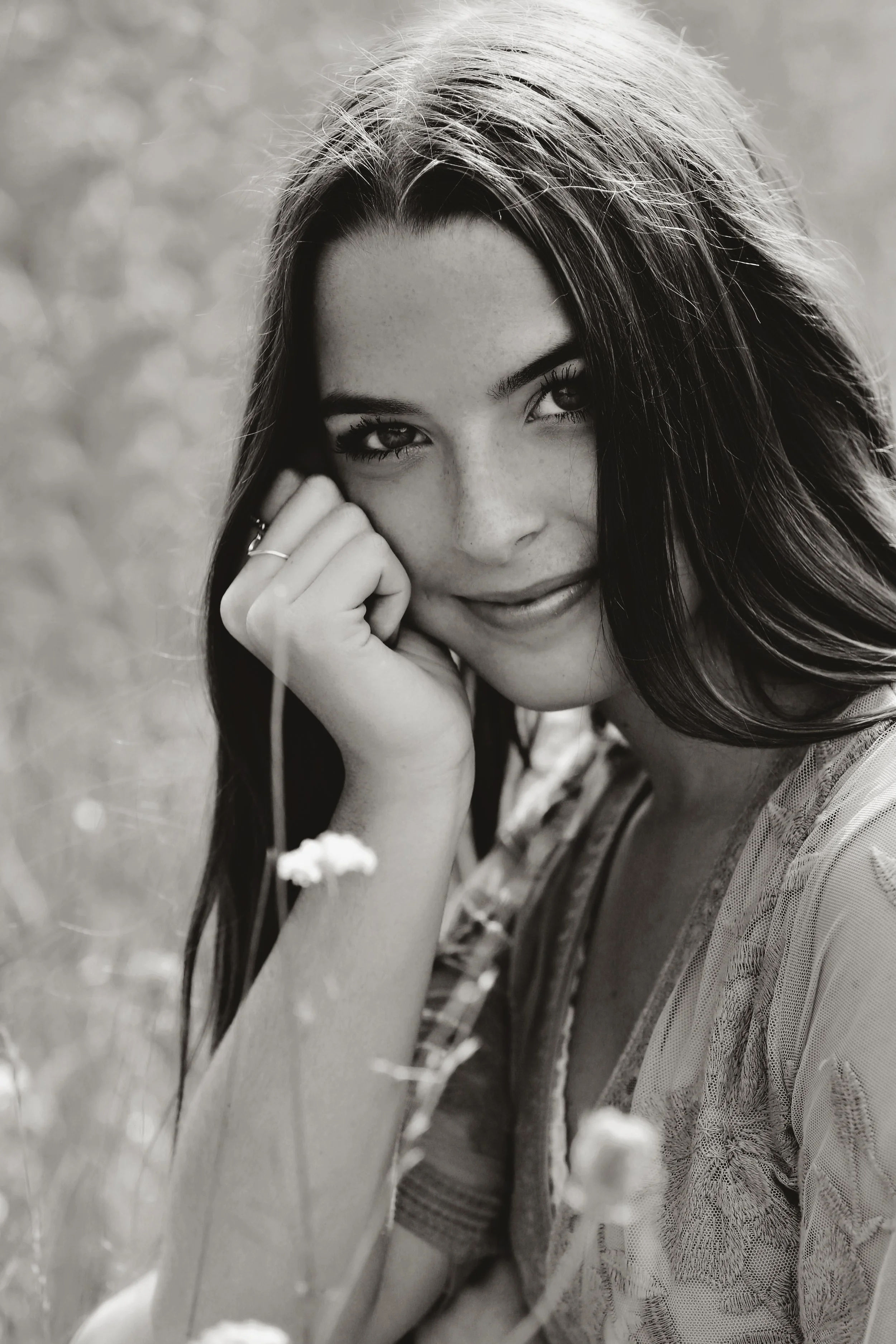 Black and white photograph of a young woman with long dark hair, smiling softly, resting her chin on her hand in a grassy outdoor setting.