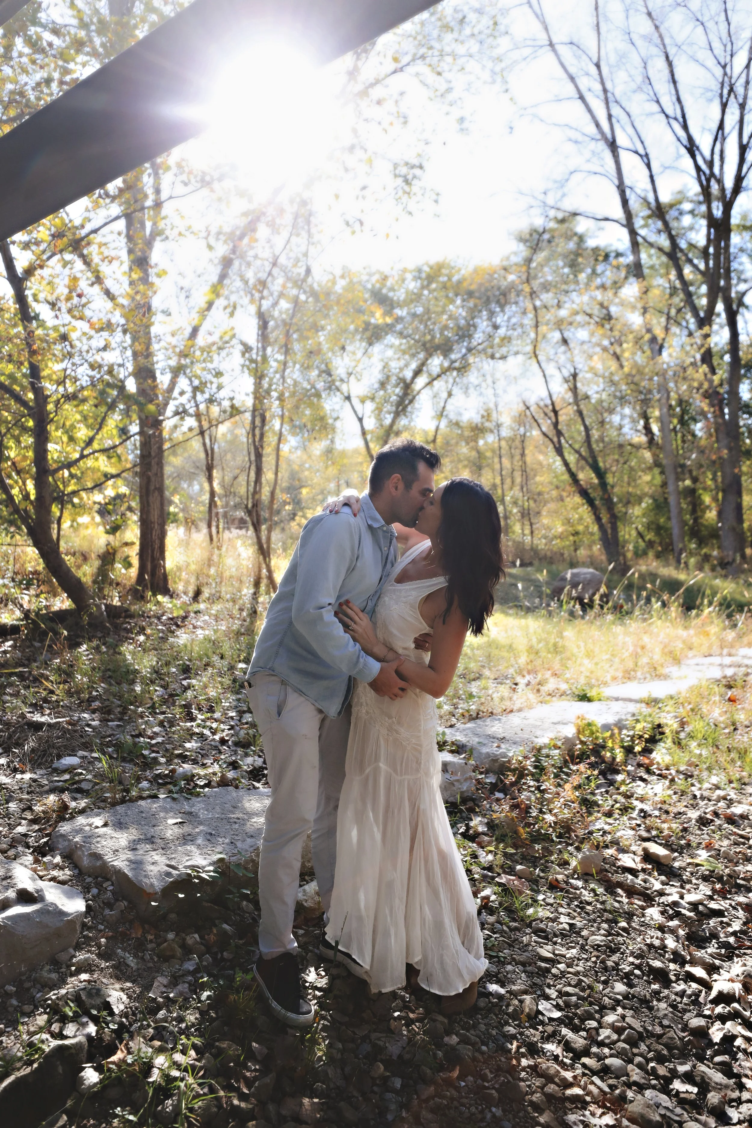 A couple kissing in a forested area underneath bright sunlight, with trees and rocks visible in the background.