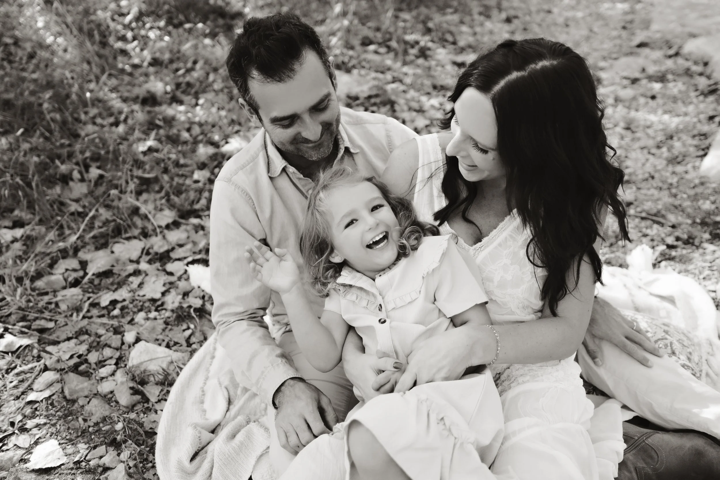 A happy family of three enjoying a moment outdoors on fallen leaves. The little girl laughs joyfully as her parents hold her and look at her lovingly.