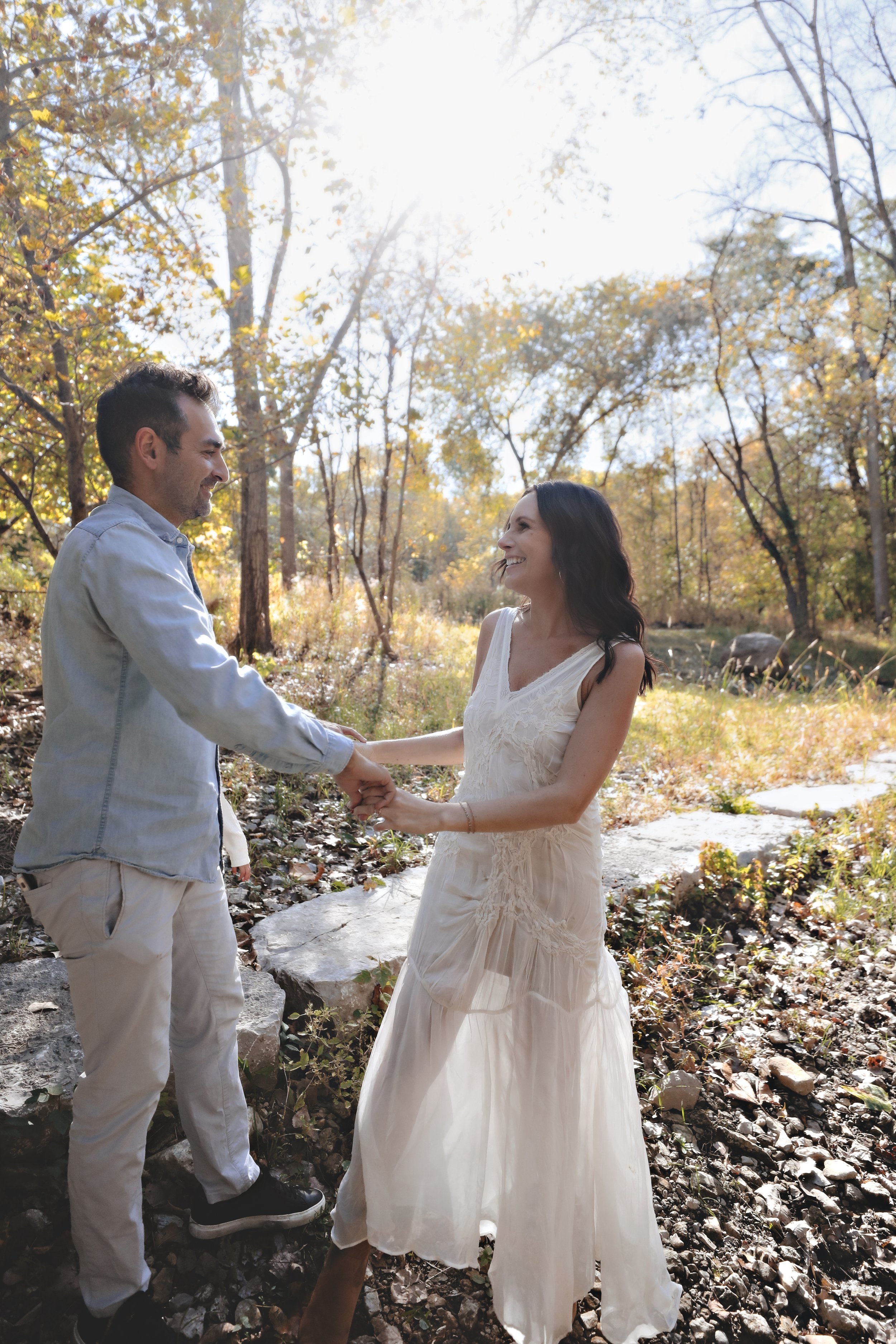 A man and a woman holding hands and smiling at each other in a wooded outdoor setting with sunlight filtering through trees.