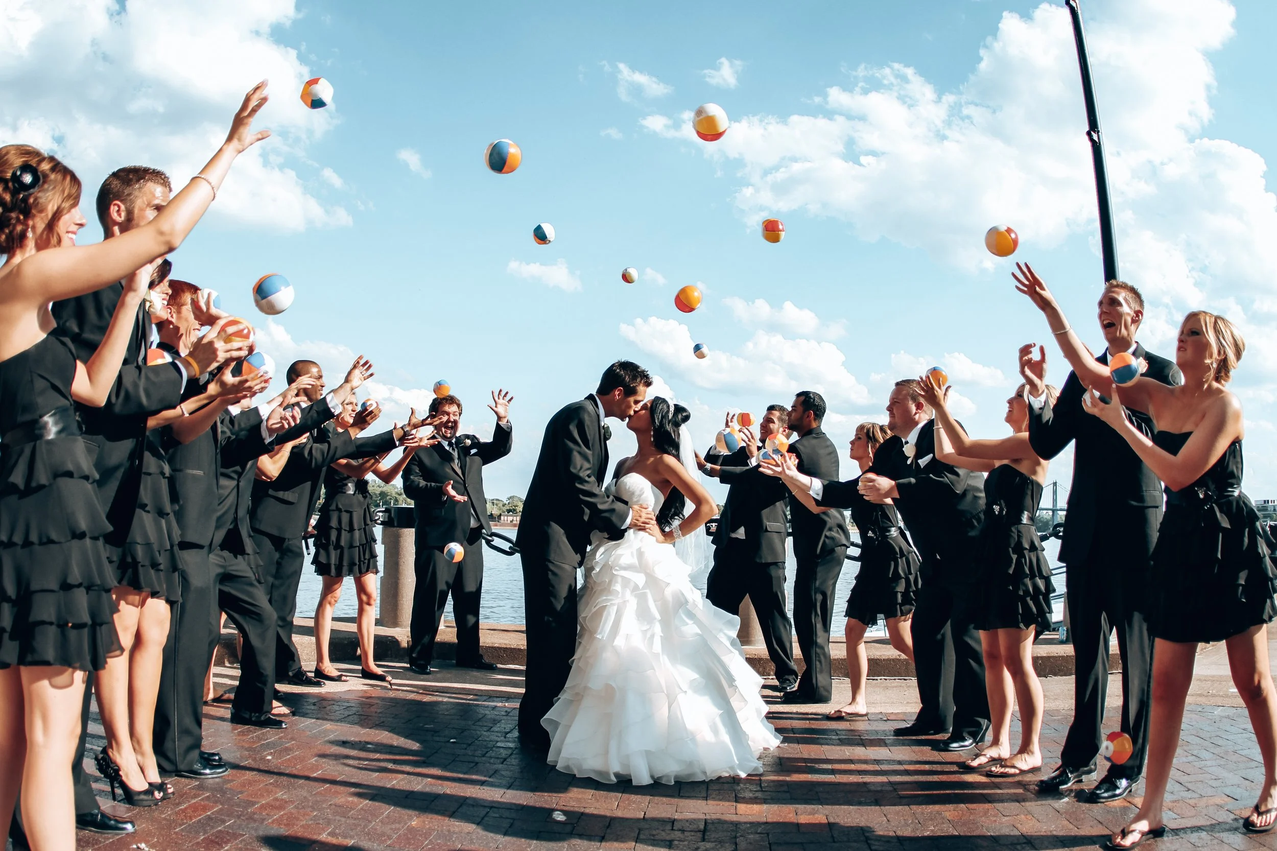 Wedding couple kissing in the center while surrounded by wedding guests throwing beach balls in the air outdoors on a sunny day.
