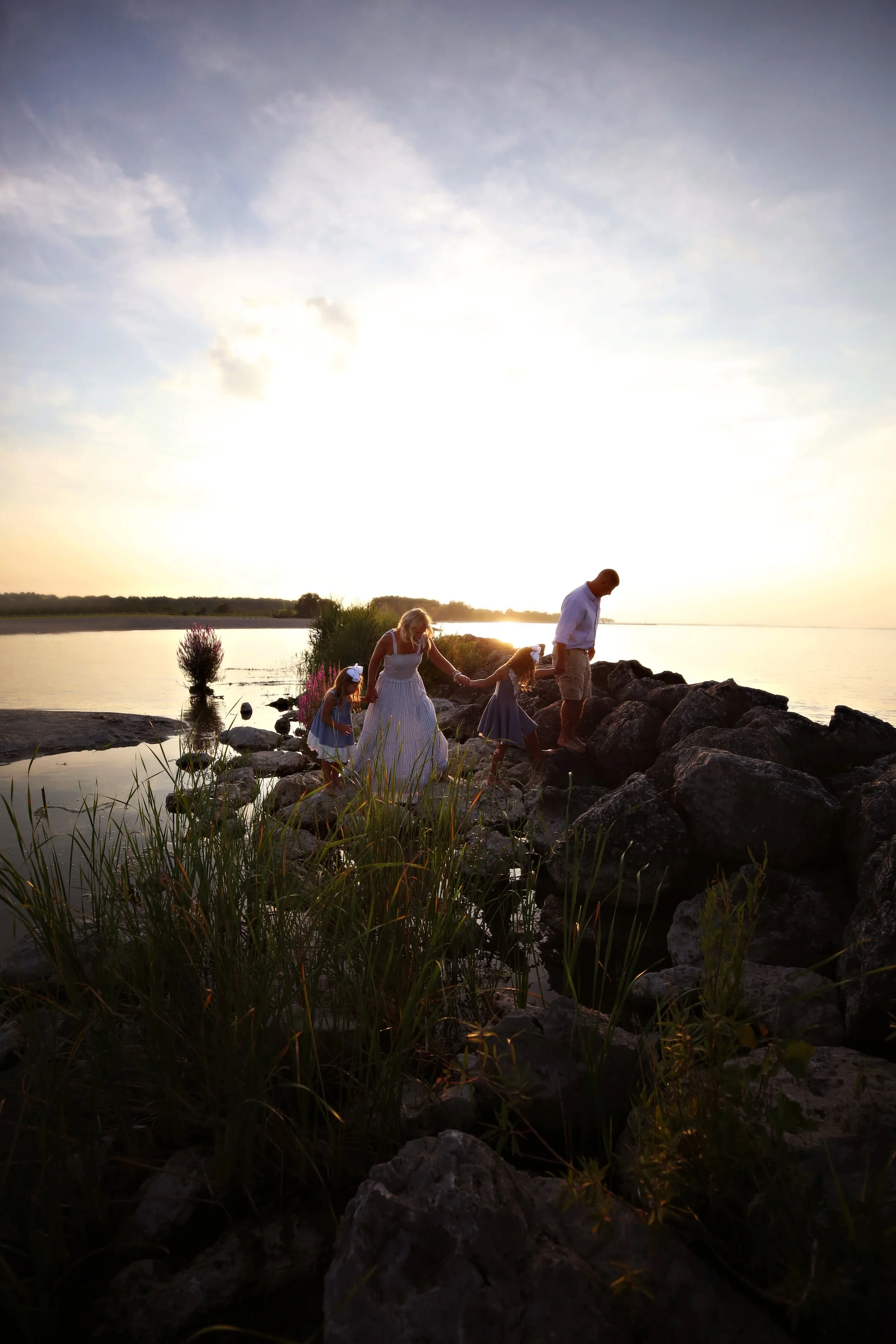 A family of four, including two young girls, walks along rocks by a calm body of water during sunset, with the sunlight creating a bright glow in the sky.