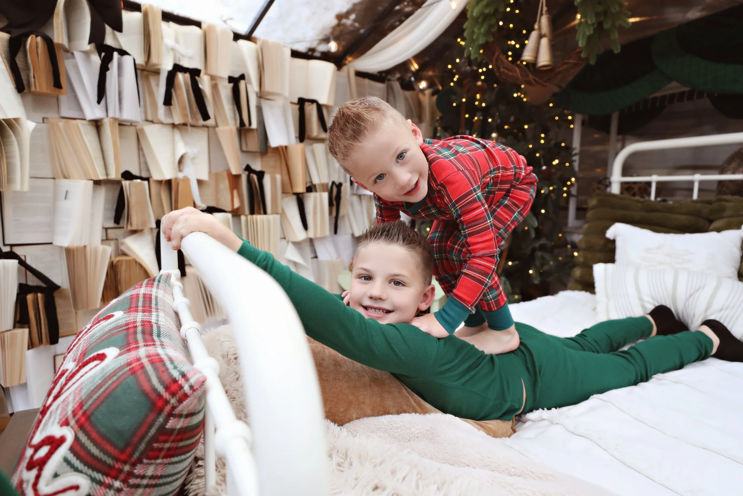 Two boys in Christmas pajamas playfully wrestling on a bed in a festive, decorated room with books, Christmas tree, and holiday decorations.