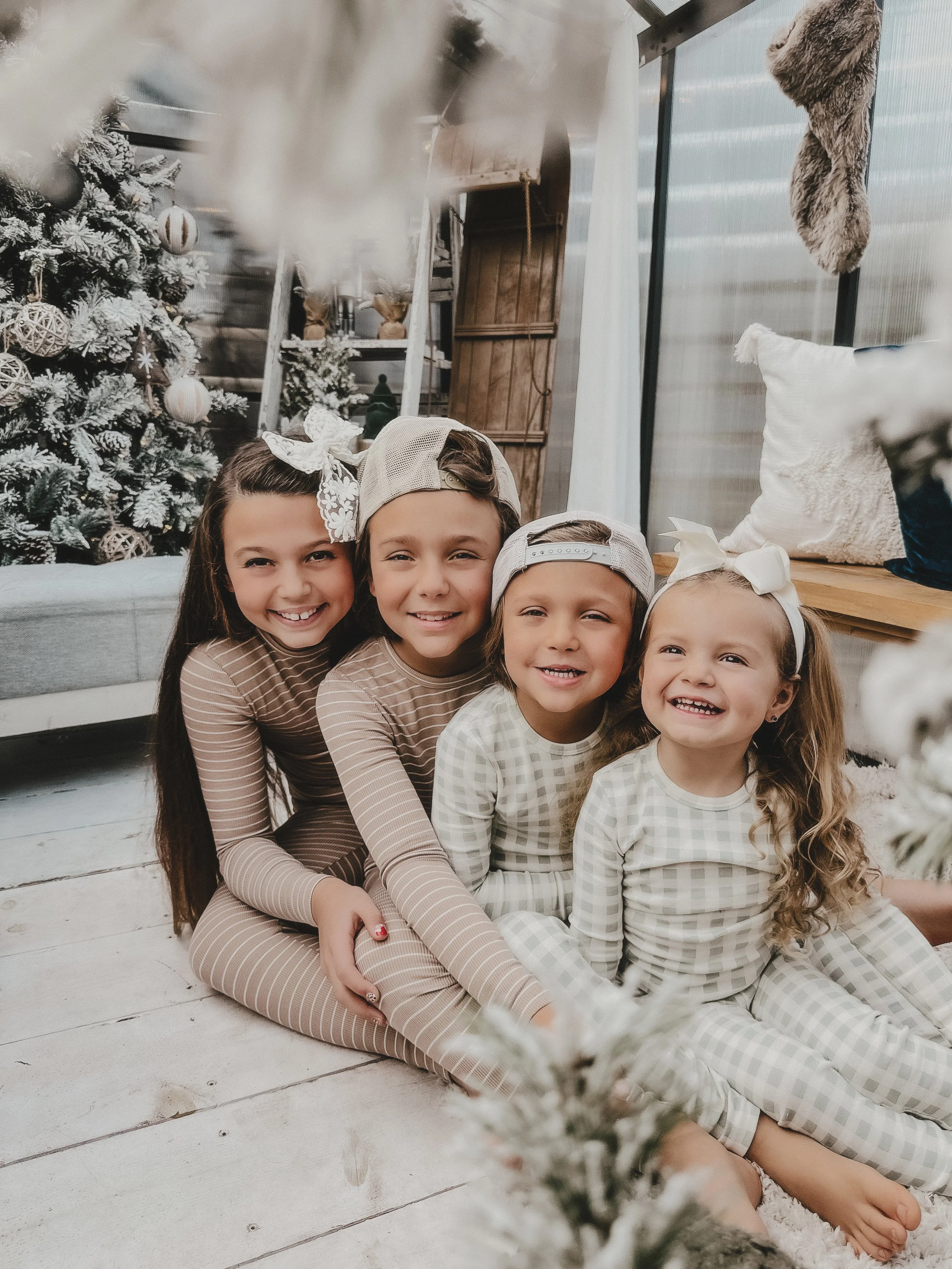 Four children sitting on the floor in front of a decorated Christmas tree, smiling at the camera.