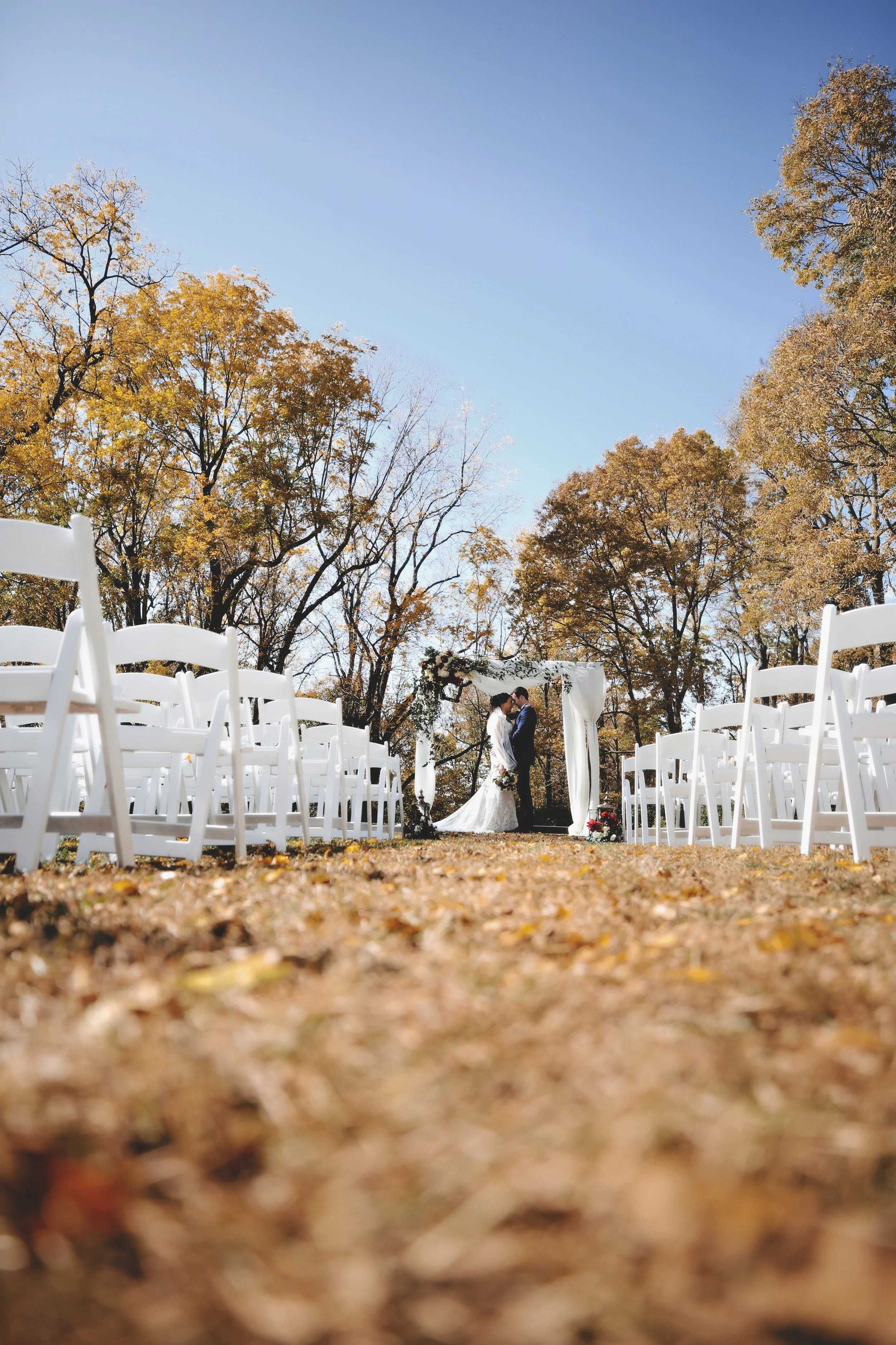 A bride and groom standing under a wedding arch outdoors during fall, with white chairs arranged on either side and a backdrop of autumn trees and a clear blue sky.