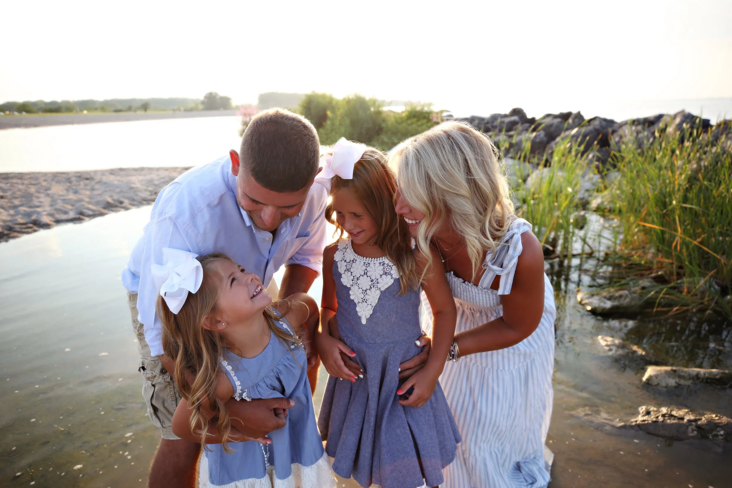 A family of four enjoying a moment by the water on a sunny day, smiling and laughing together.
