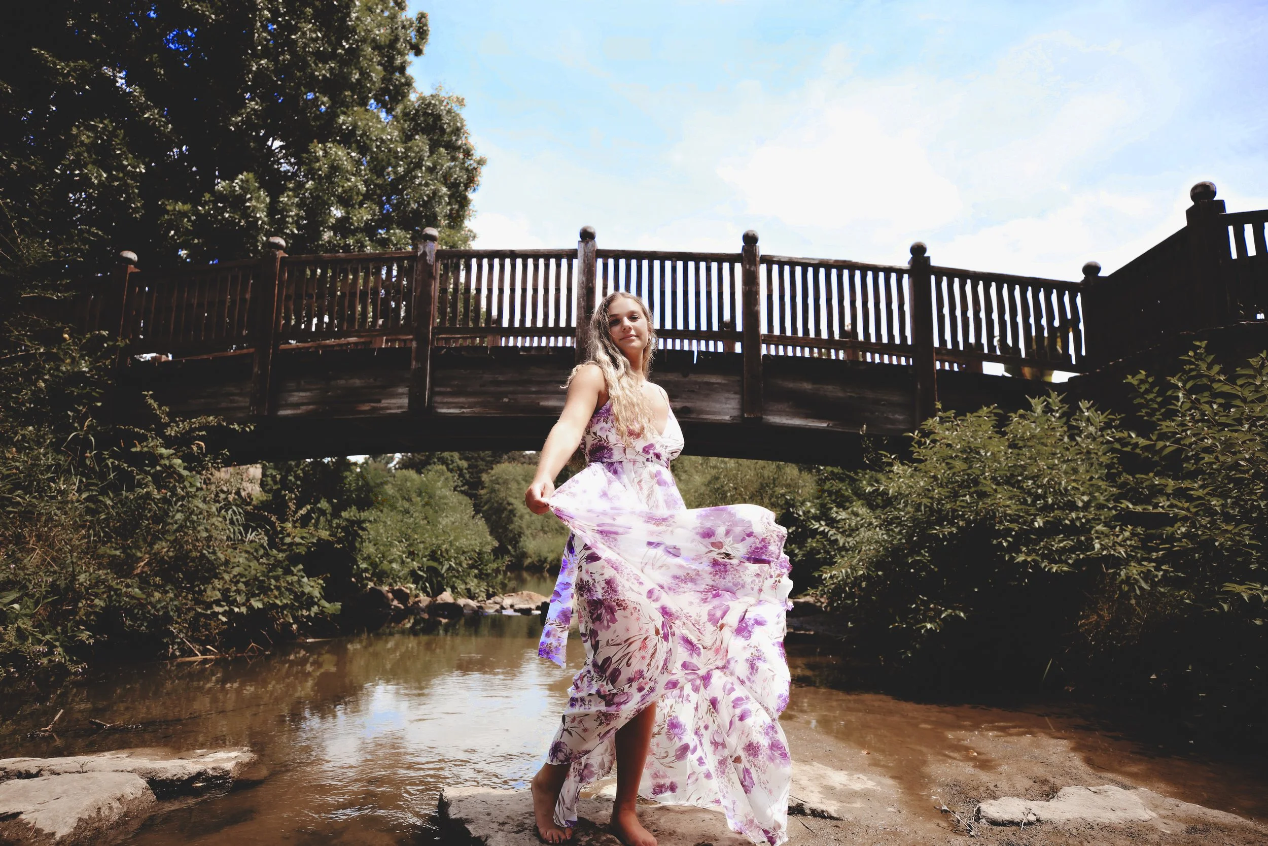A young woman in a floral dress standing in a shallow creek beneath a wooden bridge, holding up her dress as water flows around her, with trees and a partly cloudy sky in the background.