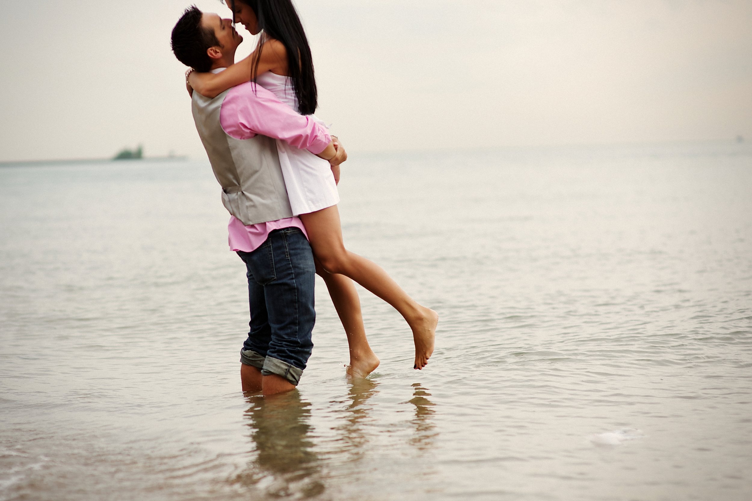 A man lifting a woman at the beach, both smiling and about to kiss, standing in shallow water with the ocean in the background.