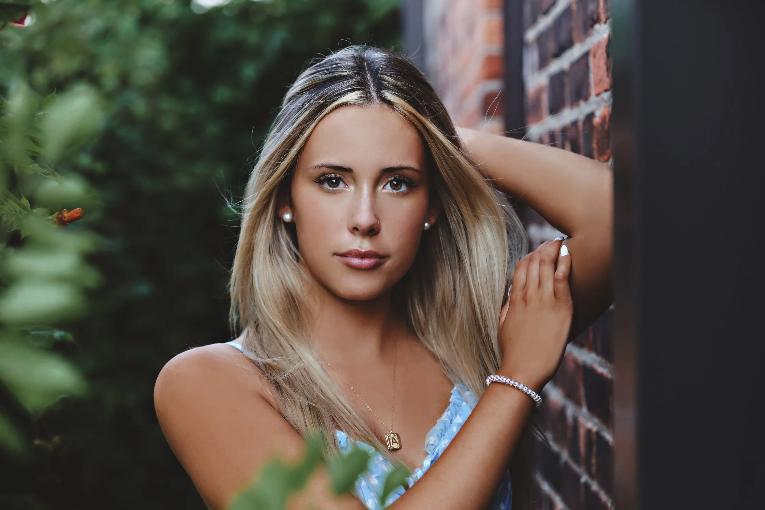 A young woman with blonde hair, wearing a blue top and jewelry, posing outdoors near a brick wall with greenery in the background.