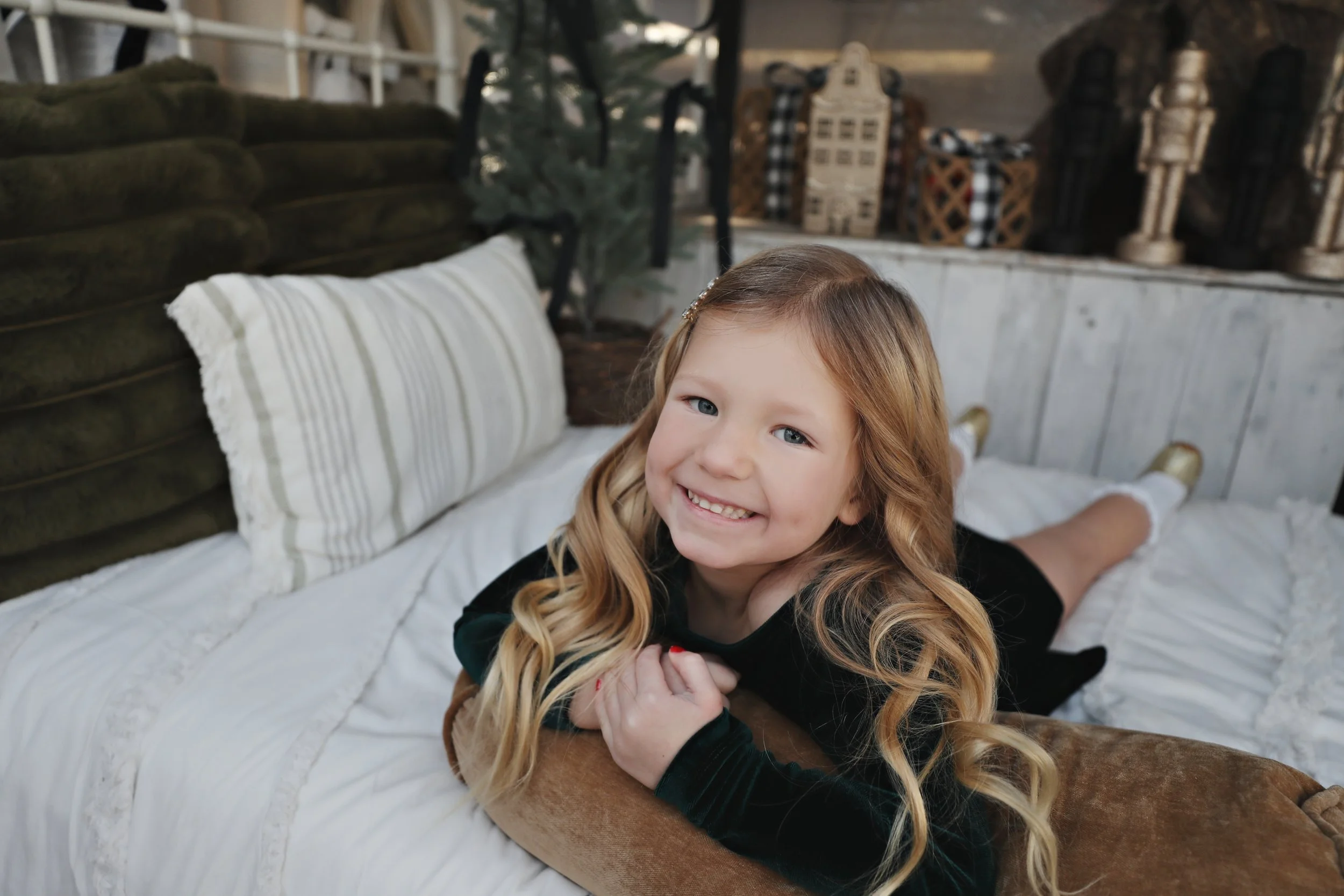 A young girl with long blonde hair and a big smile lying on her stomach on a bed with white linens and striped pillow, looking at the camera.