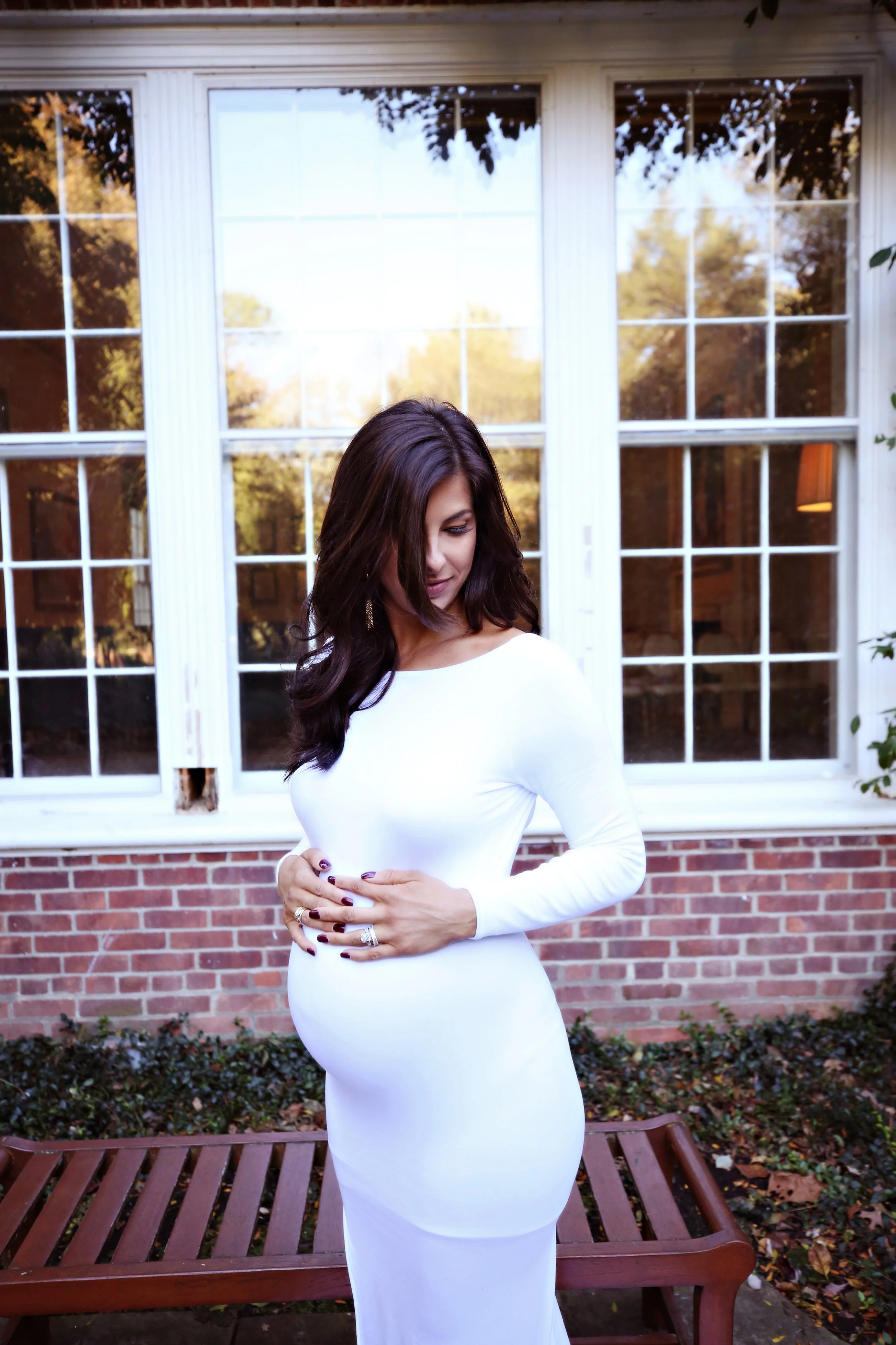 A woman in a fitted white long sleeve dress standing outdoors in front of a house with large windows and brick facade, looking down and to her left, with her hands clasped on her stomach.